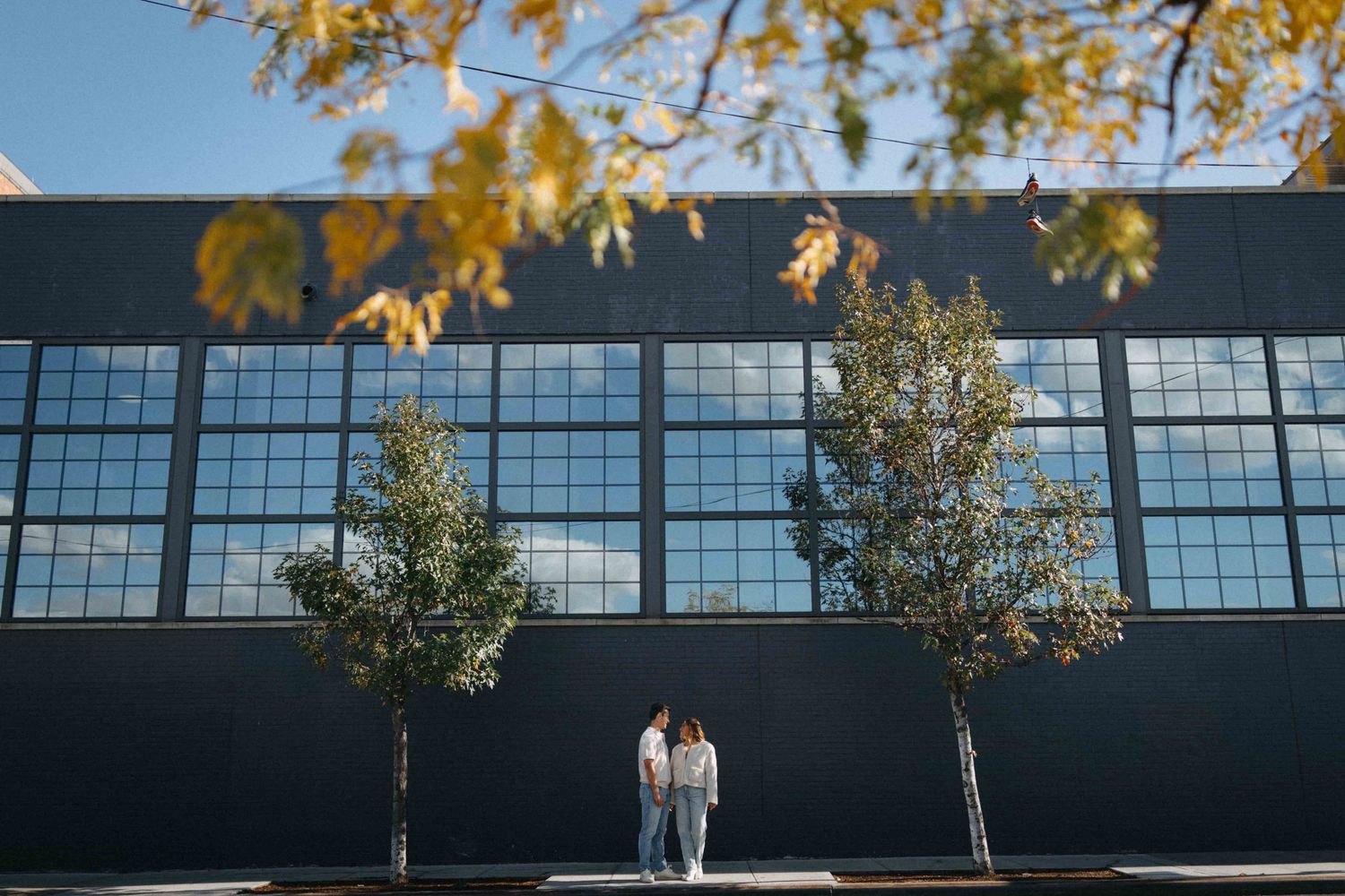 A couple stands together in front of a large industrial building with autumn trees framing the scene.