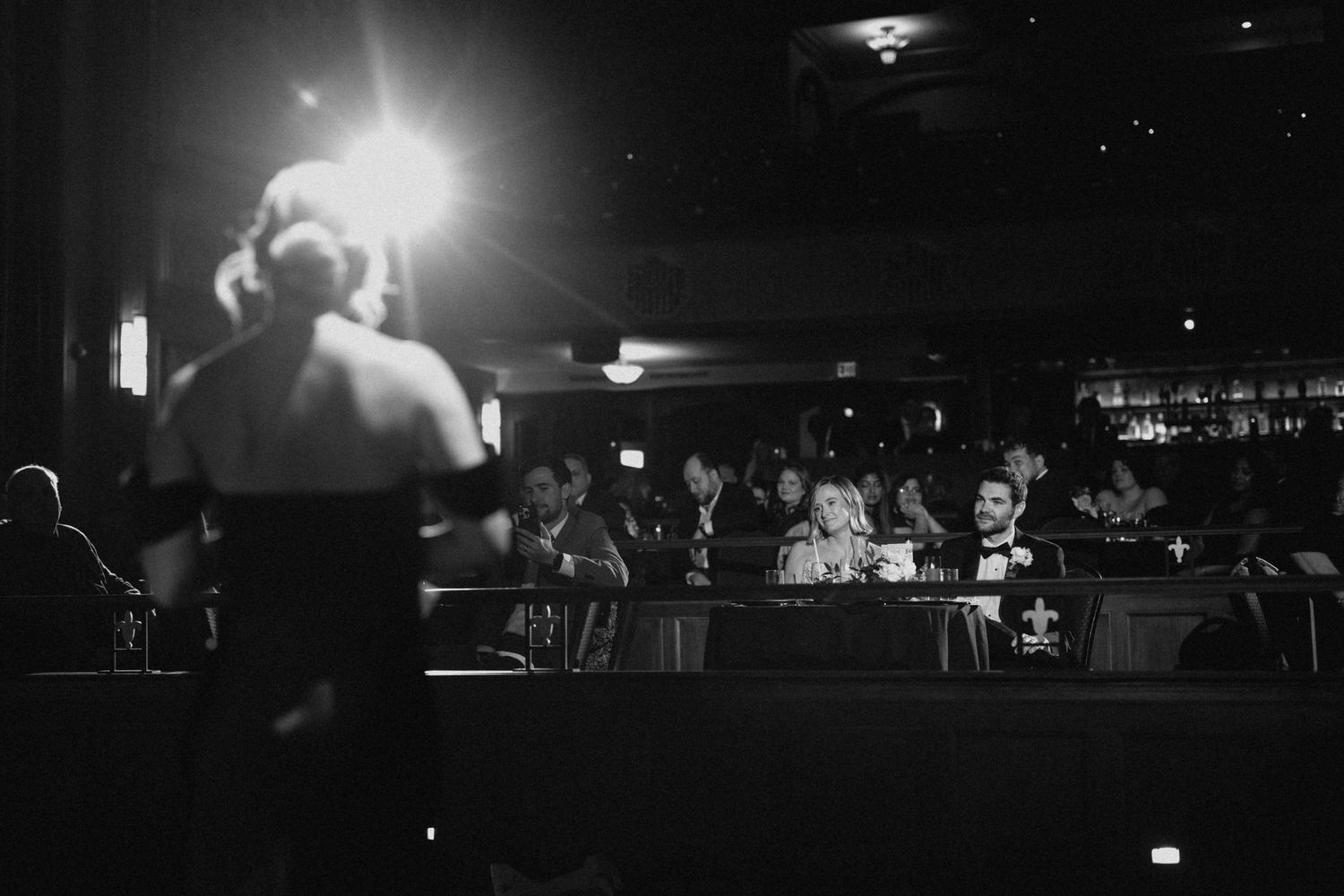 Black and white photo of a performer on stage entertaining seated wedding guests in a dimly lit venue.