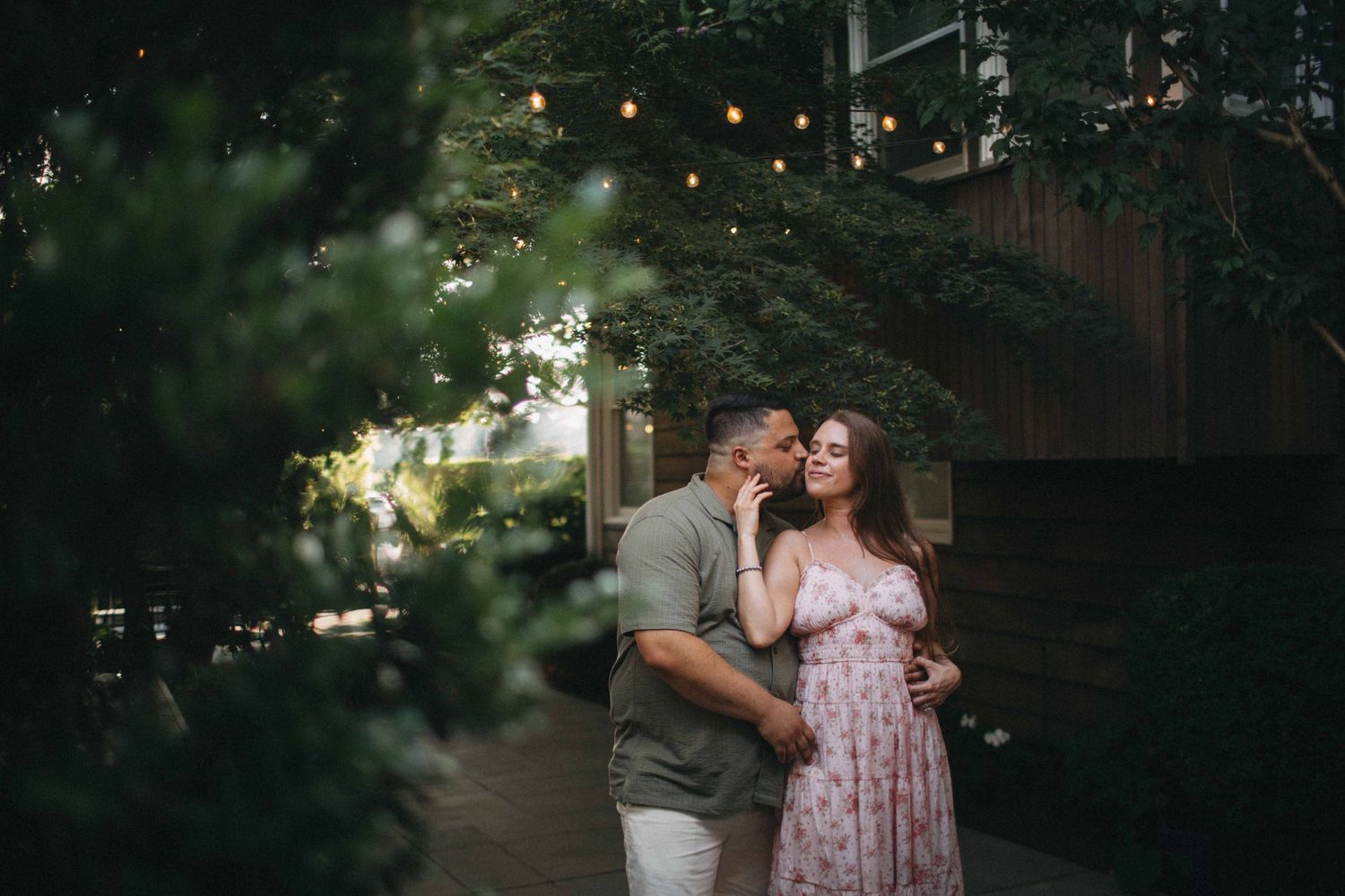 Couple sharing a tender kiss surrounded by lush greenery and string lights at dusk.