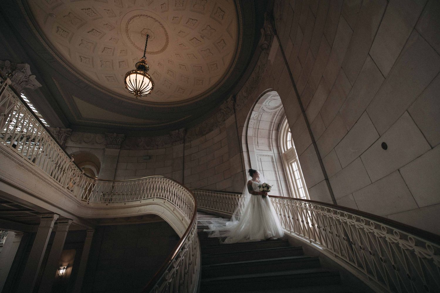 Bride in flowing white gown stands on an ornate curved staircase beneath a grand chandelier in an elegant venue.