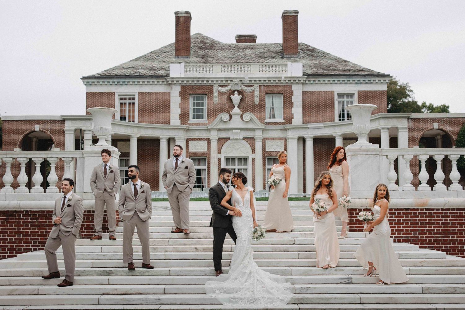 Wedding party posing on grand white steps in front of a classic brick mansion estate venue.