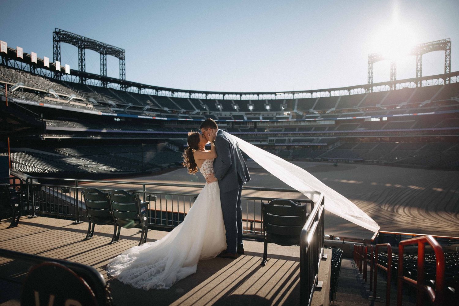 Bride and groom share a kiss on a sun-drenched terrace with a baseball stadium in the background.