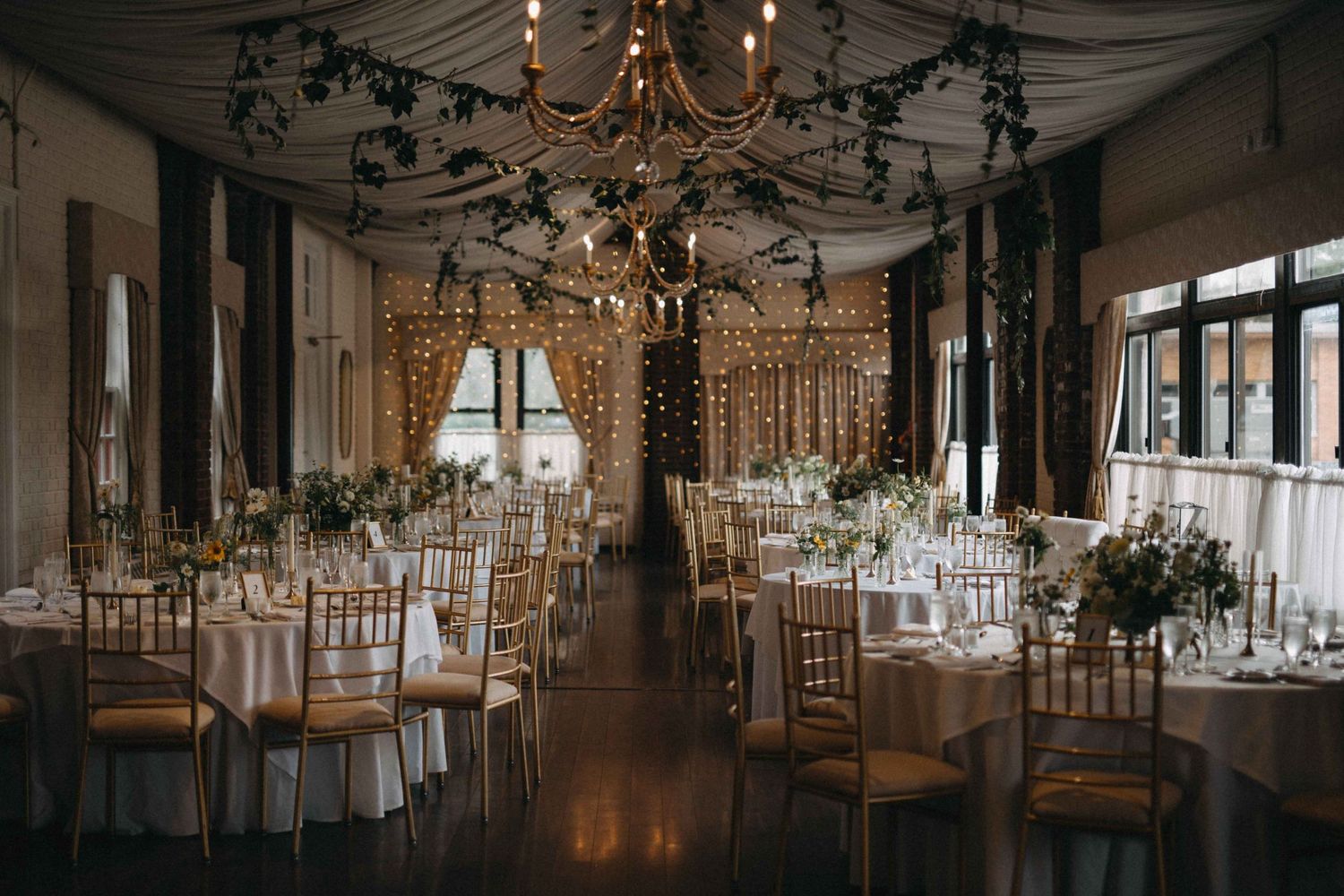 Romantic wedding reception hall with chandelier, string lights, and floral centerpieces on long tables.