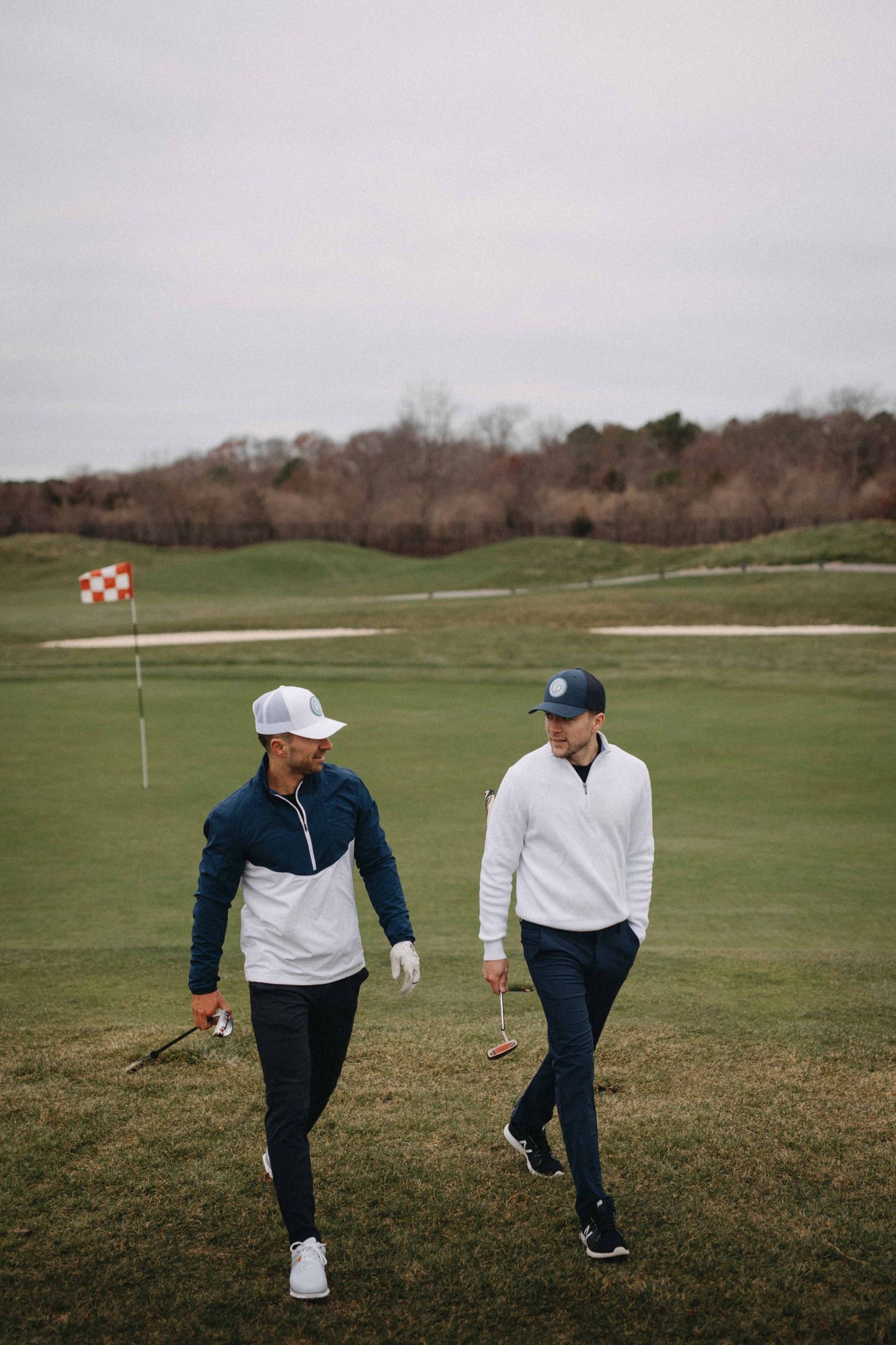 Two men in golf attire walking on a green golf course with a flag and sand bunker in the background.