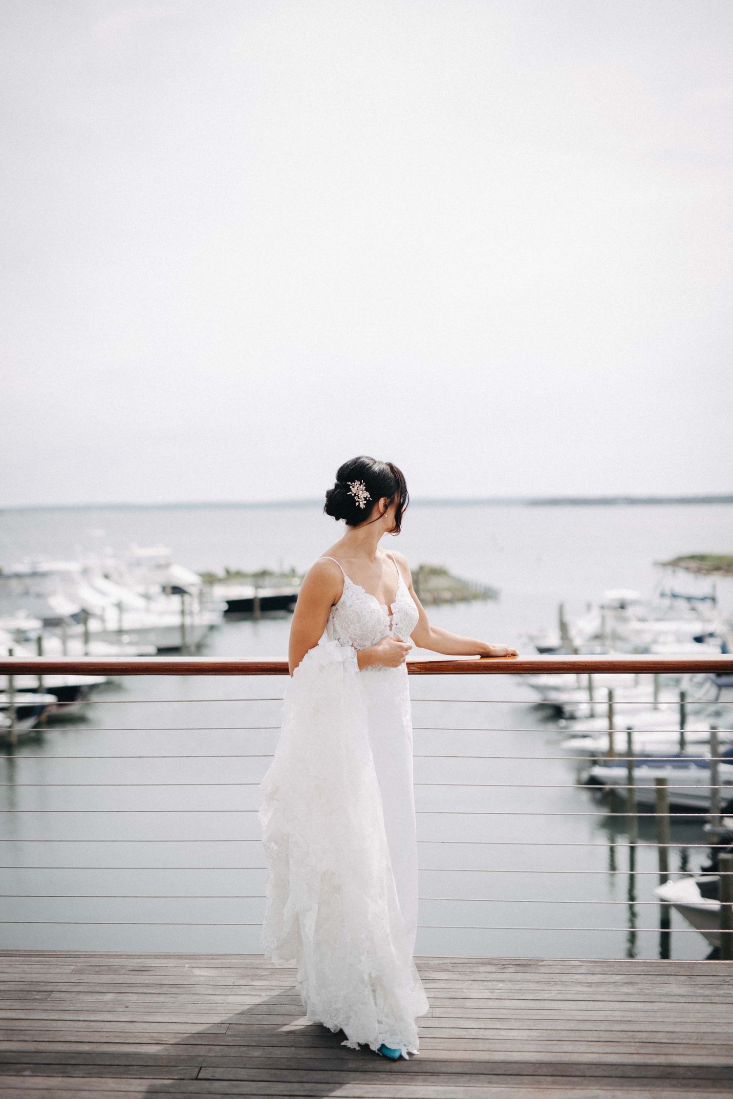 Bride in a flowing white lace wedding gown standing on a marina deck overlooking calm harbor waters.