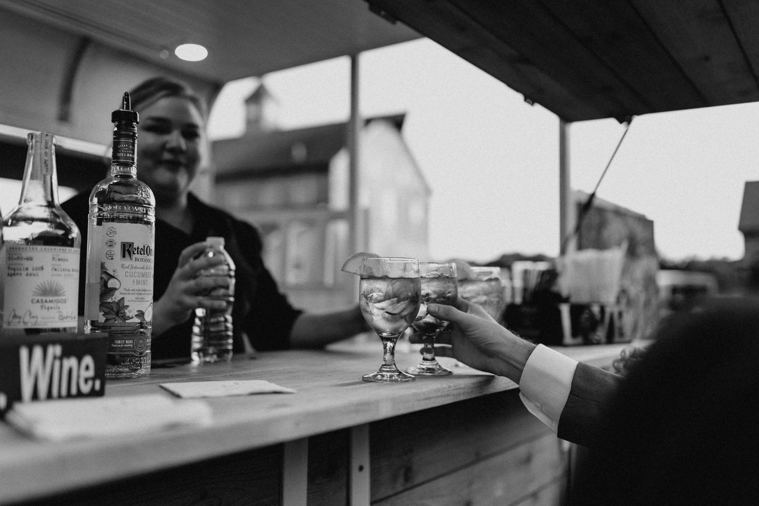 Black and white photo of bartender serving drinks at outdoor wedding bar with wine sign visible.