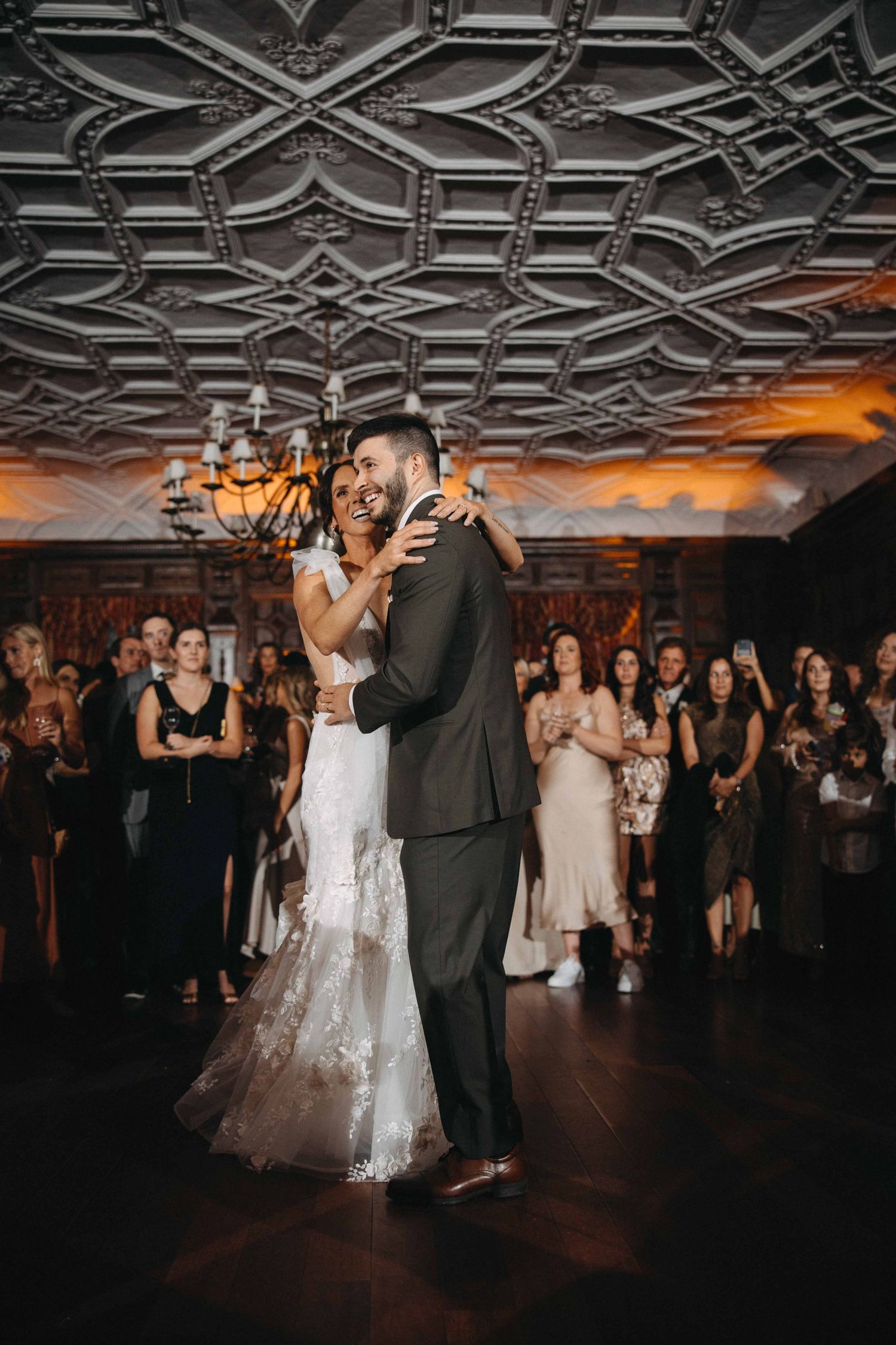 Bride and groom share a joyful first dance under an ornate ceiling with chandelier while guests applaud around them.