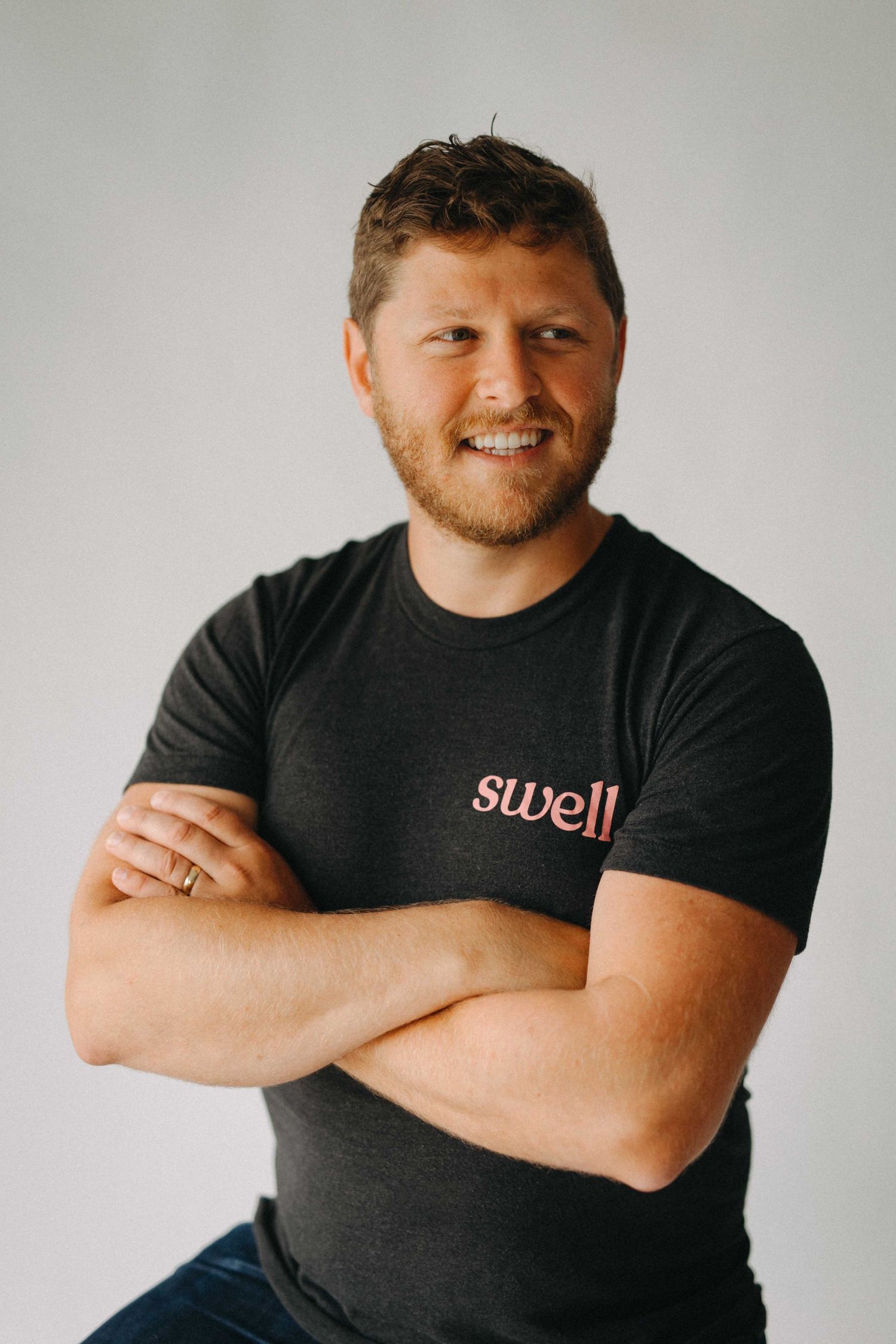 Man in dark Swell branded t-shirt smiling with arms crossed against light gray background.