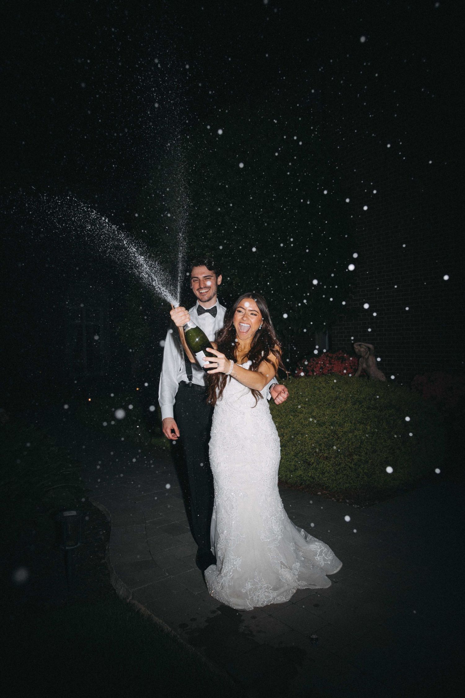 Joyful newlyweds spraying champagne bottle outdoors at night celebrating their wedding reception.