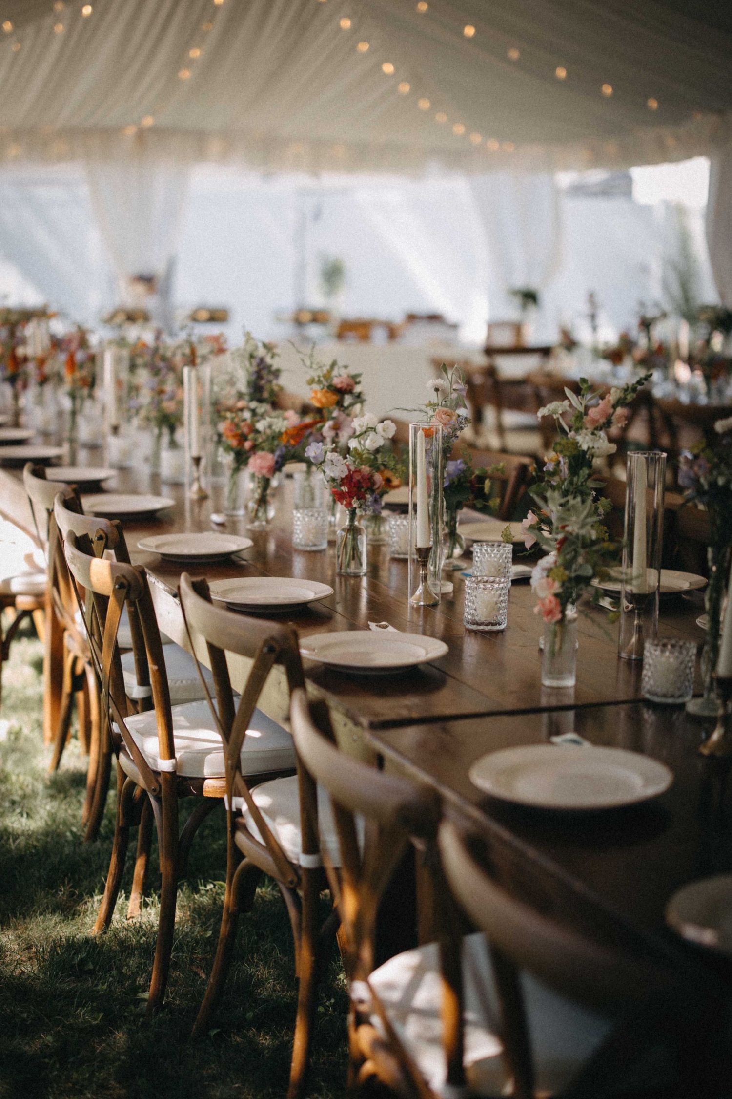 Rustic outdoor wedding reception with wooden cross-back chairs and colorful floral centerpieces under tent.
