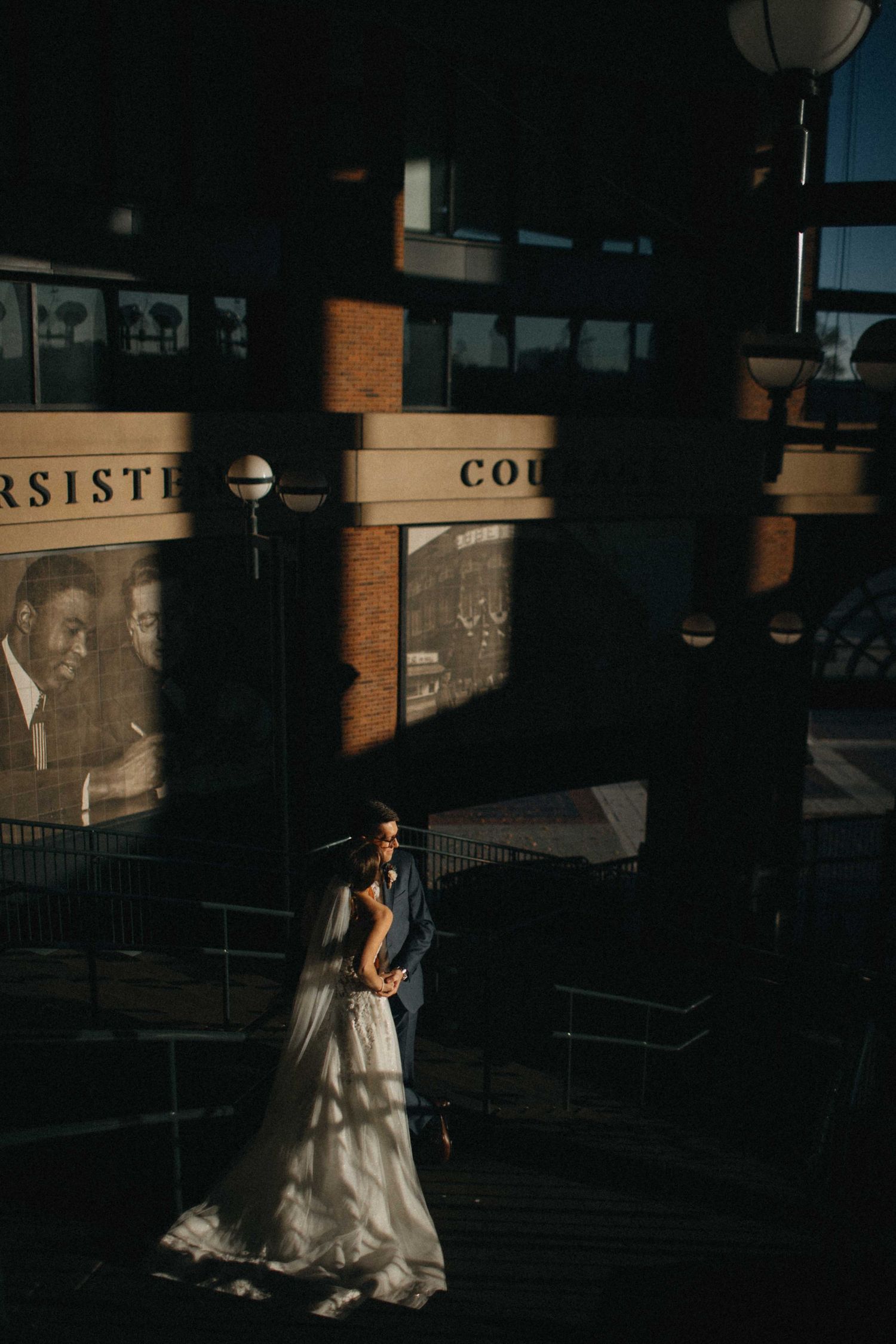 Bride and groom embracing in dramatic shadows and golden light outside an urban brick building.