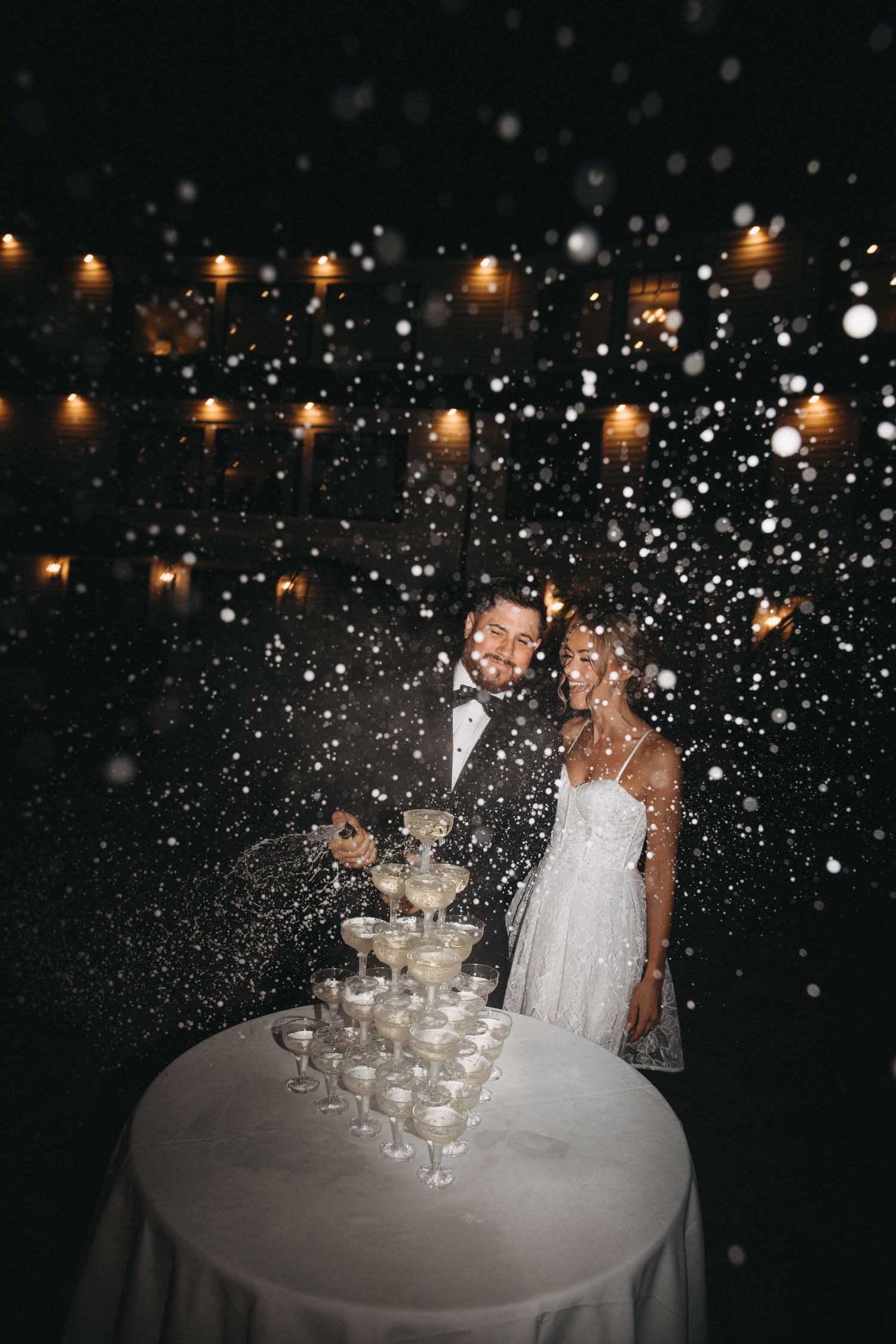 Bride and groom celebrate champagne tower pour with sparkling confetti falling at night reception.