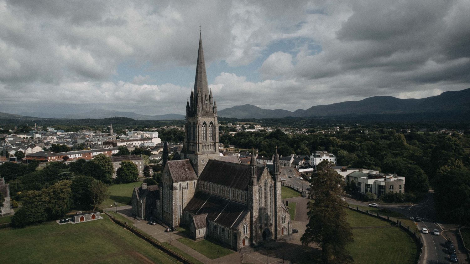 Aerial drone view of a Gothic church with tall spire surrounded by lush greenery and mountain backdrop.