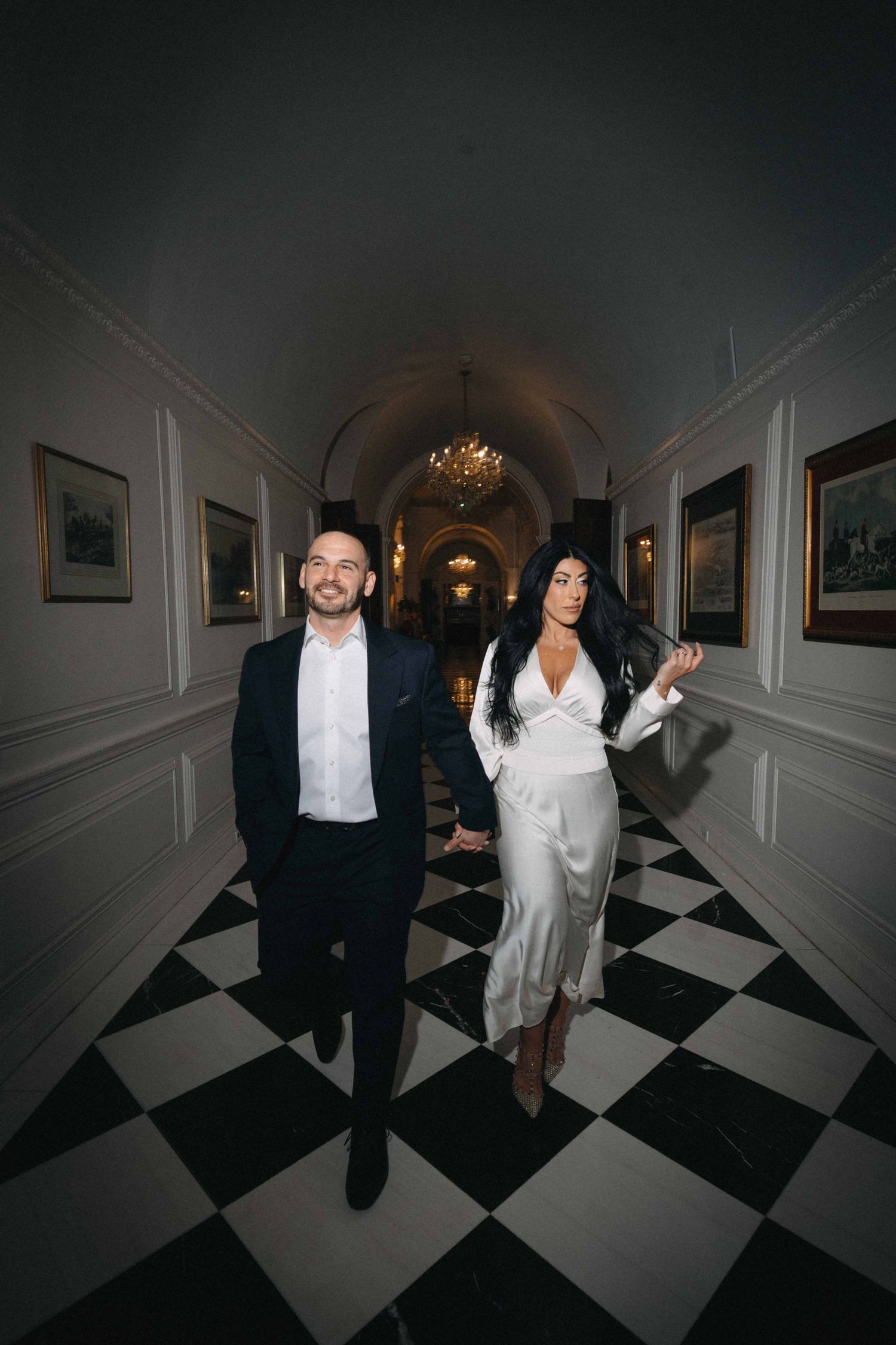 Elegant couple walking hand-in-hand through a grand hallway with checkered floors and arched ceiling.