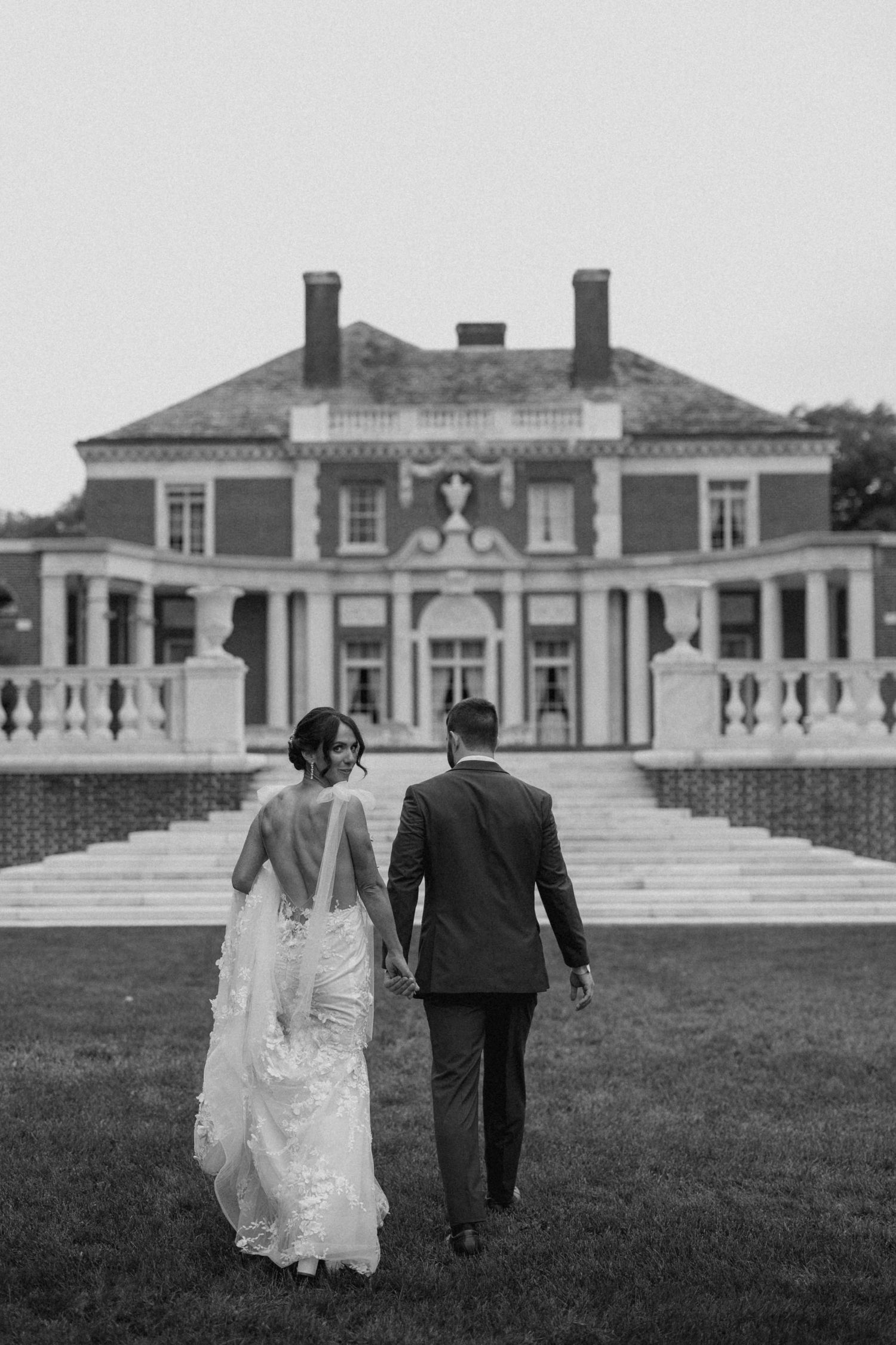 Black and white photo of bride and groom walking hand in hand toward an elegant mansion wedding venue.