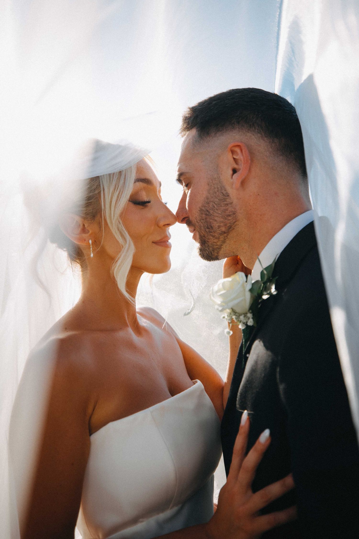 Bride and groom share intimate moment under sheer veil in soft backlit sunlight.