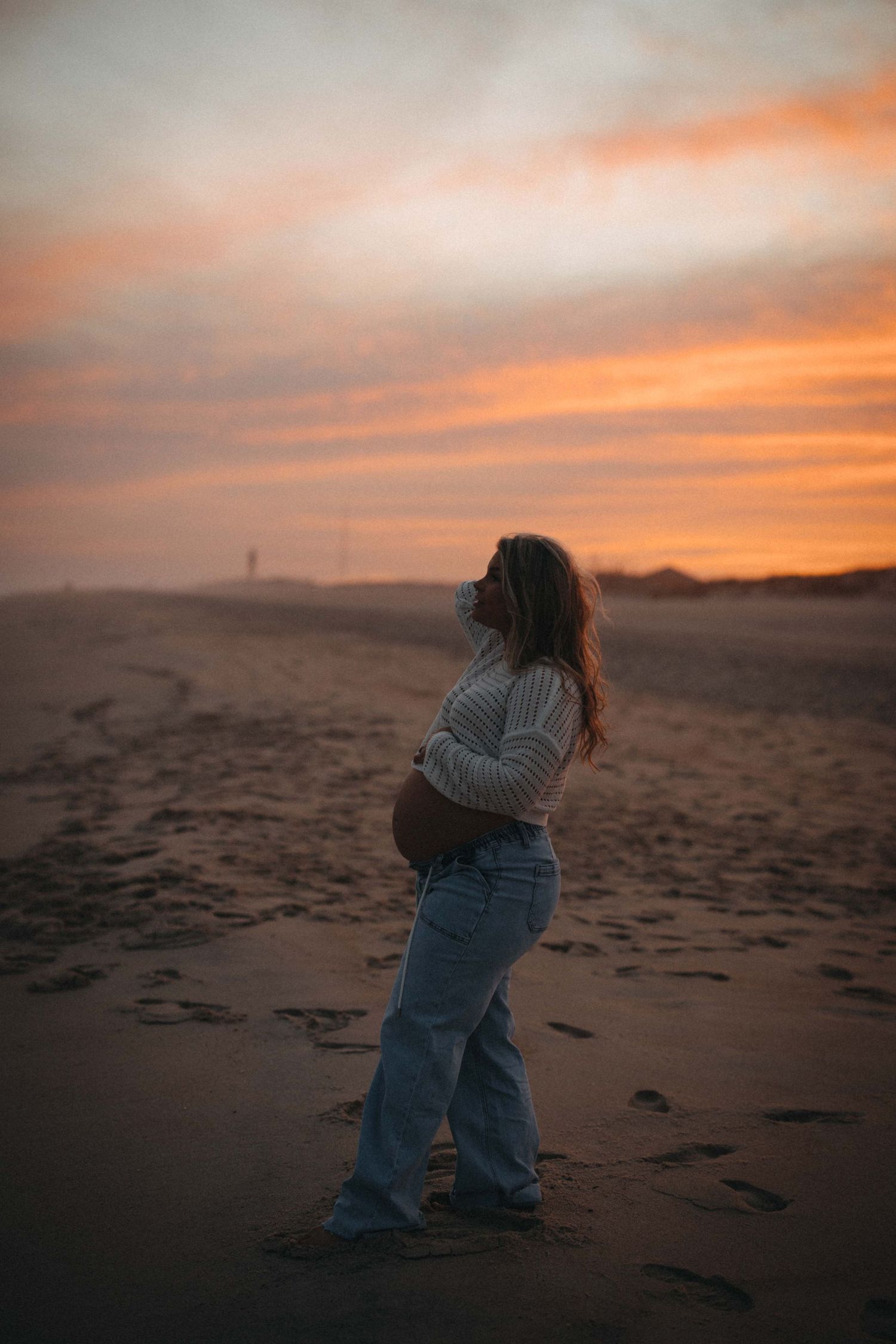 A pregnant woman cradles her belly on a sandy beach during a stunning orange and pink sunset.