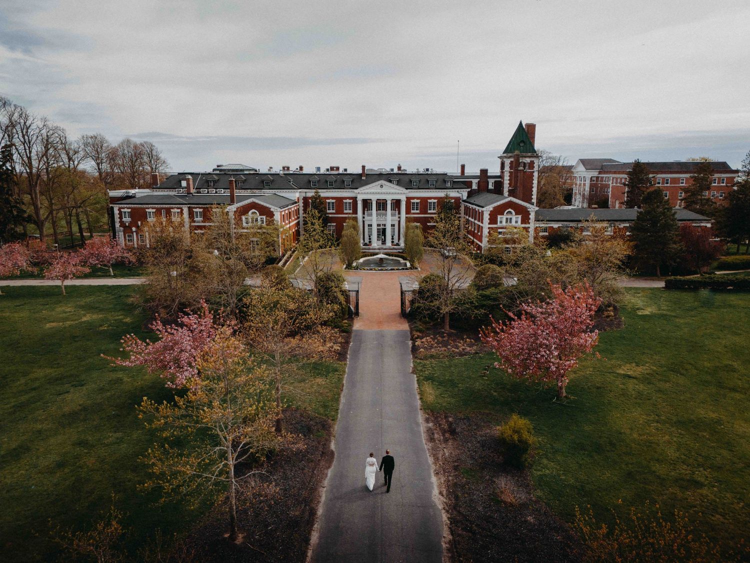 Aerial drone view of bride and groom walking path toward elegant brick building with spring blossoms.