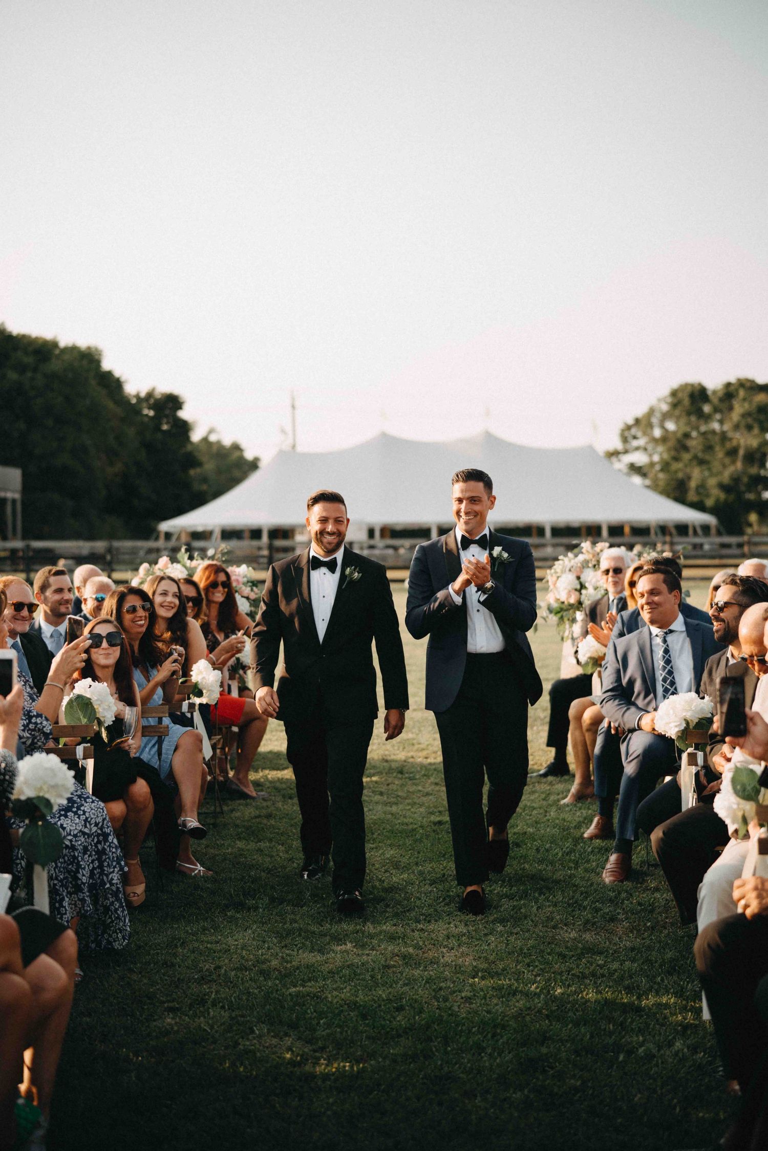 Two grooms in tuxedos walk hand in hand down aisle after same-sex wedding ceremony outdoors.