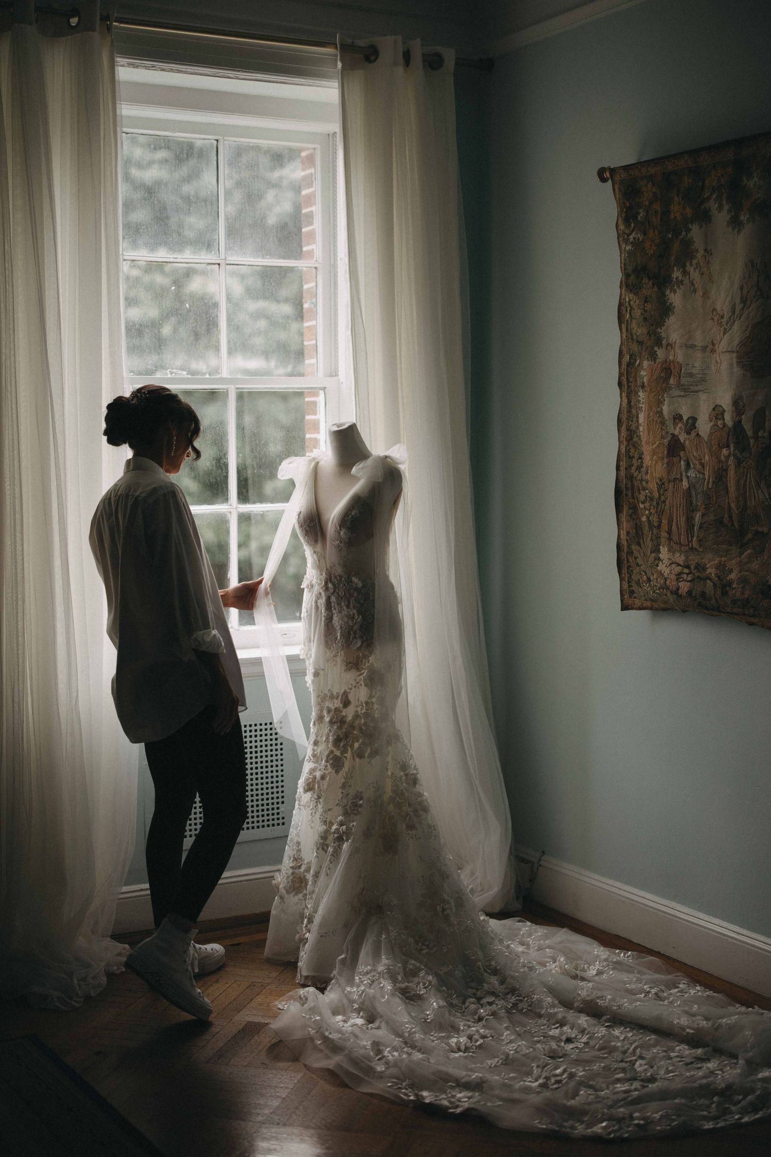 Bridal stylist adjusting an ornate lace wedding gown on a mannequin by a bright window in an elegant room.