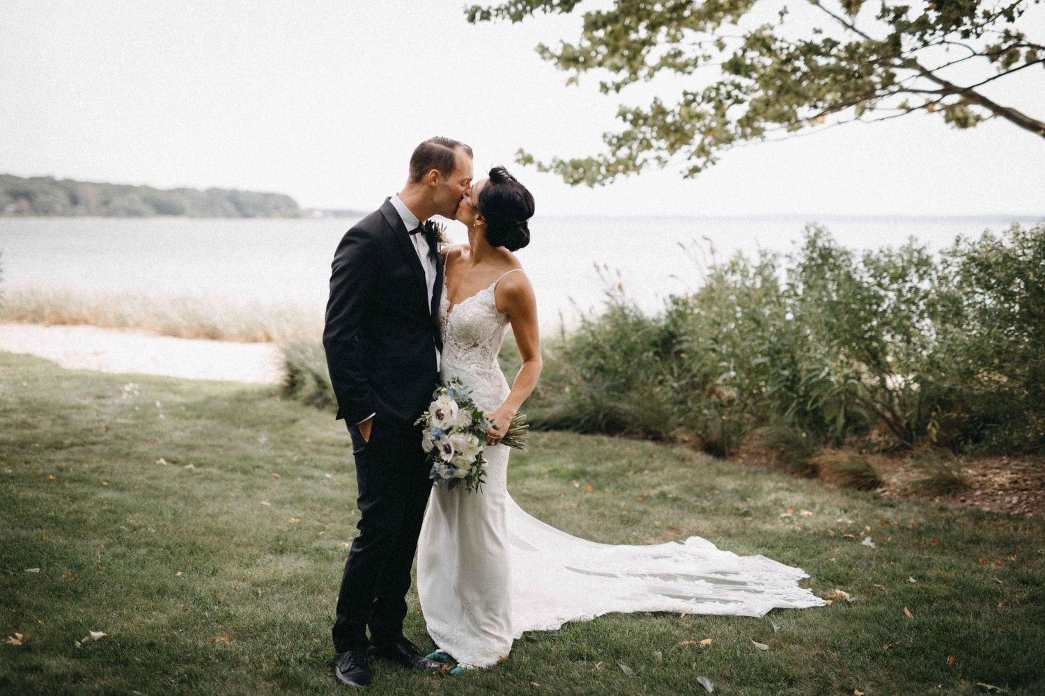 Bride and groom sharing a romantic kiss on a grassy lawn with a serene waterfront backdrop.