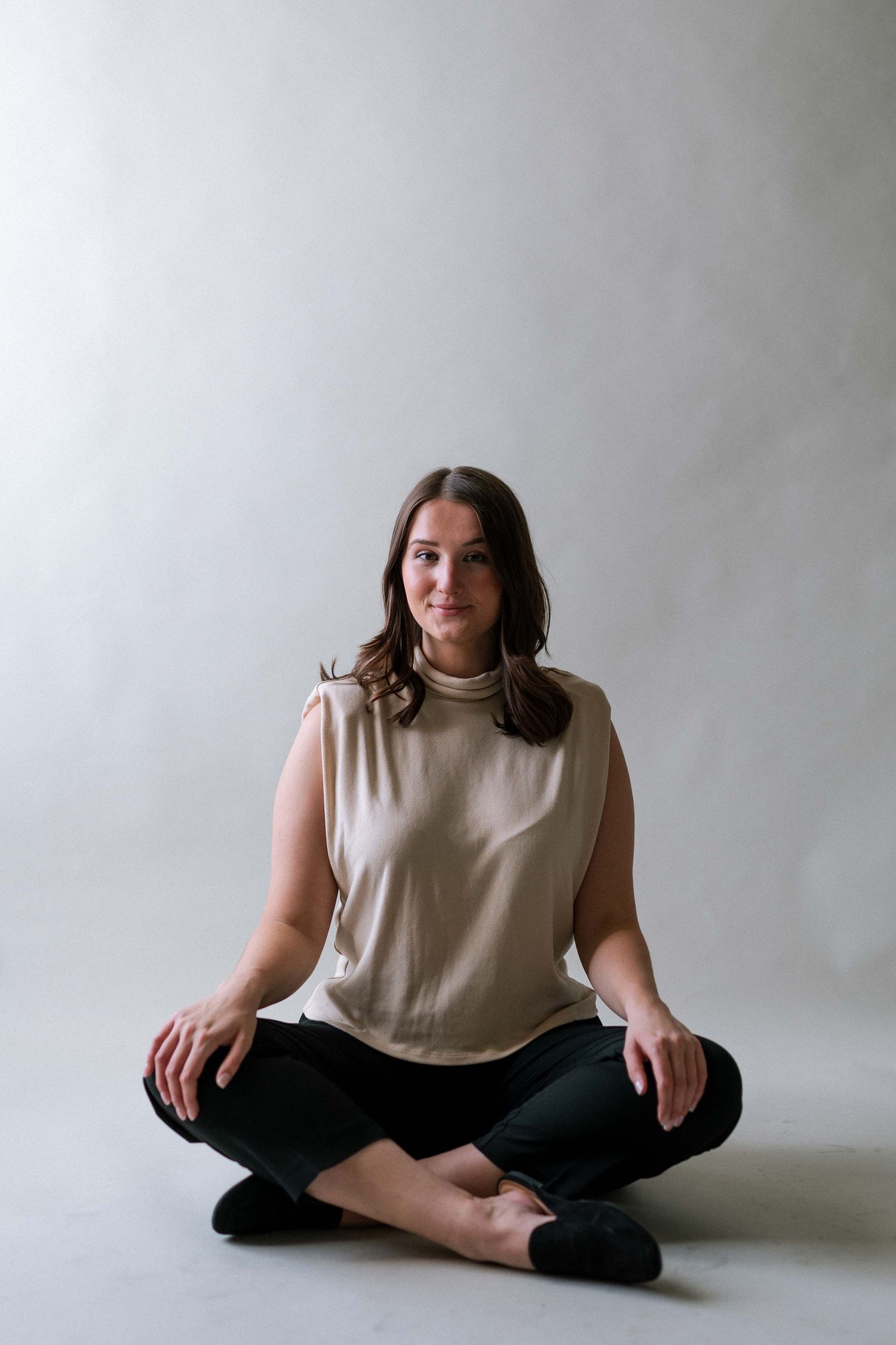 Woman in beige sleeveless top and black pants sitting cross-legged on the floor against a gray wall.