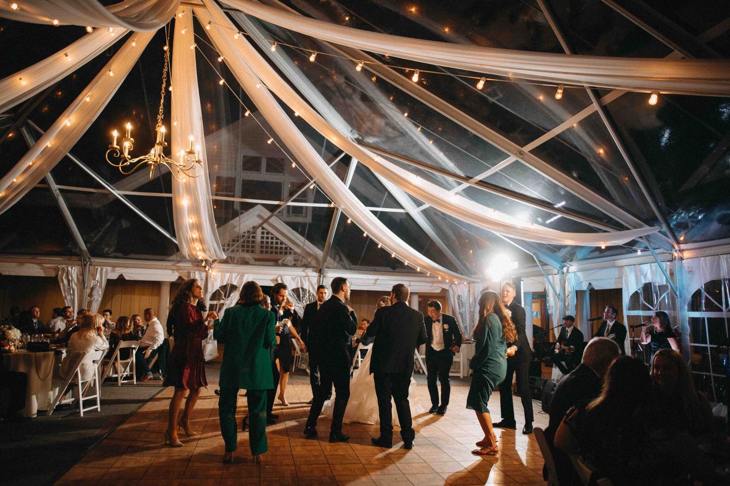 Guests dance under draped fabric and string lights inside elegant clear wedding reception tent at night.