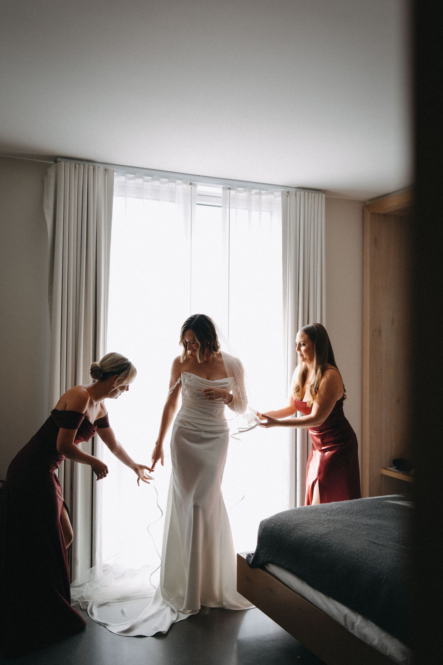 Two bridesmaids in red dresses helping bride into elegant white fitted wedding gown by window.