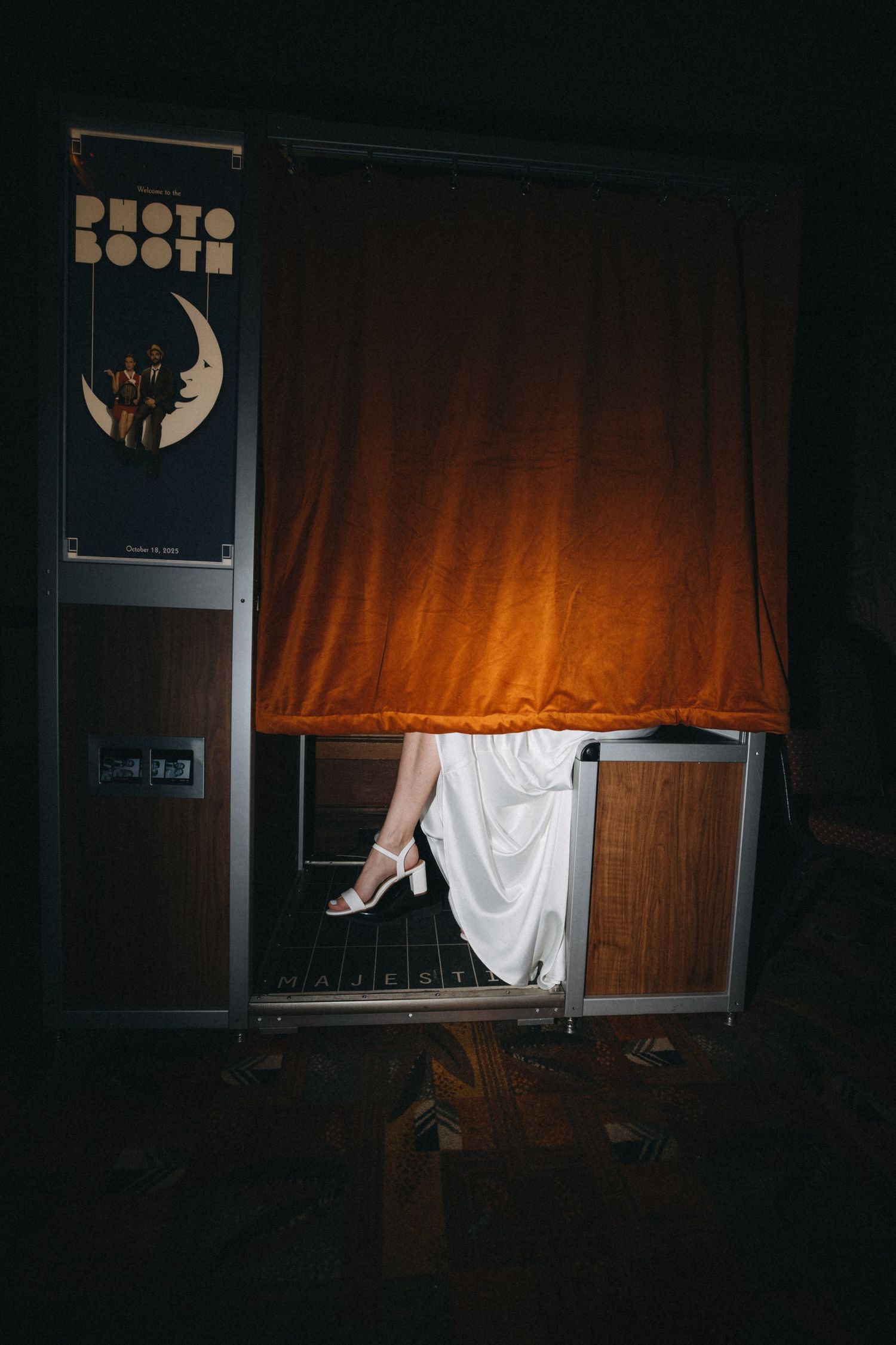 Bride in white dress and heels peeks out of vintage photo booth with orange velvet curtain at wedding.