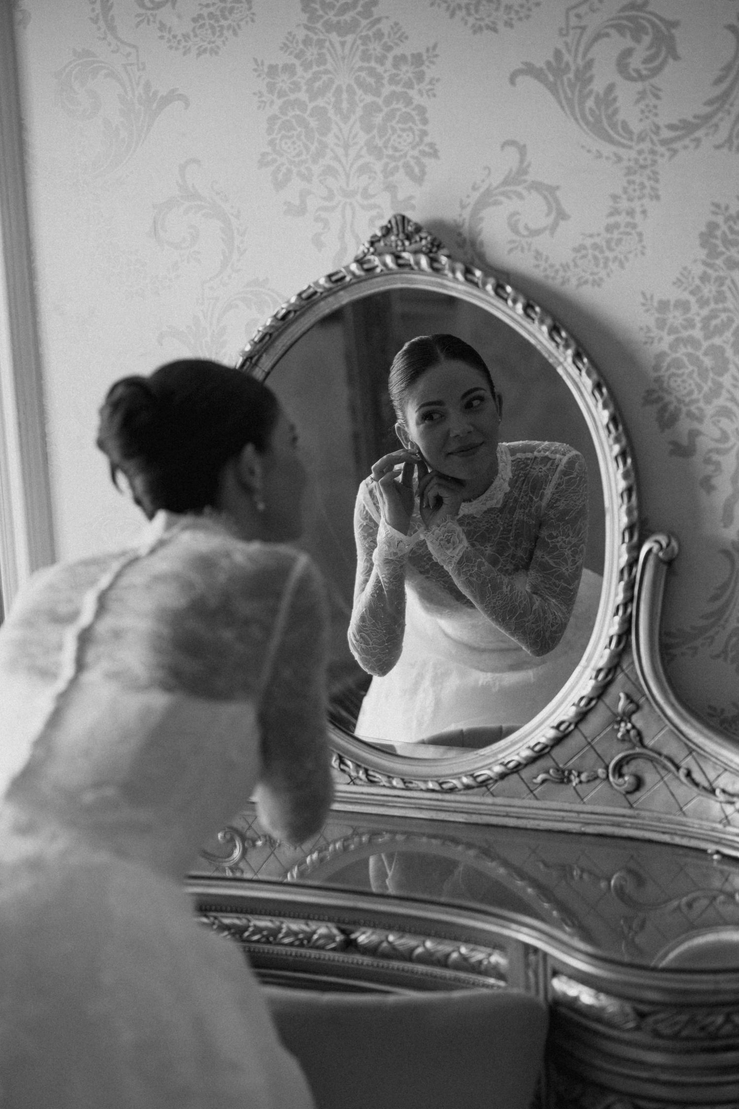 Bride in white lace dress adjusting earring before ornate vintage mirror in black and white wedding photo.