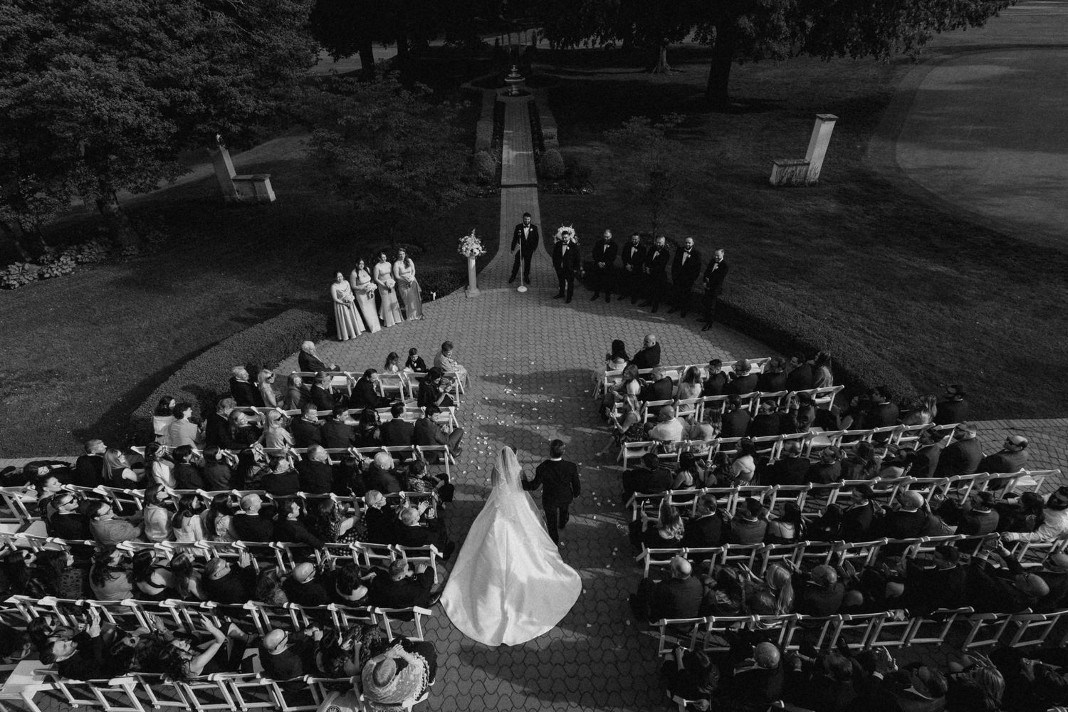 Aerial black and white photo of an outdoor wedding ceremony with bride, groom, wedding party, and seated guests on a lawn.