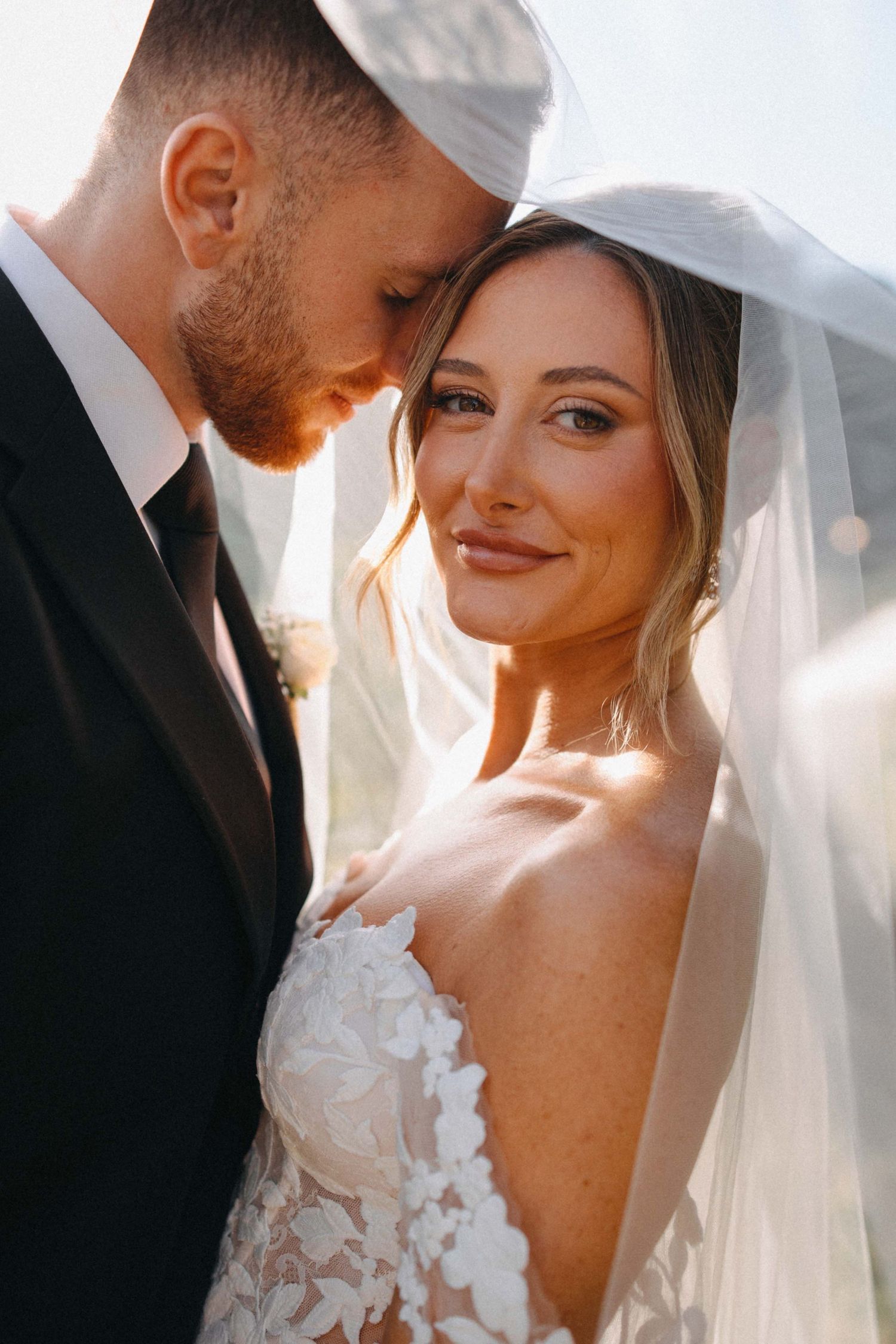 Bride and groom sharing intimate moment under sheer veil in golden sunlight.