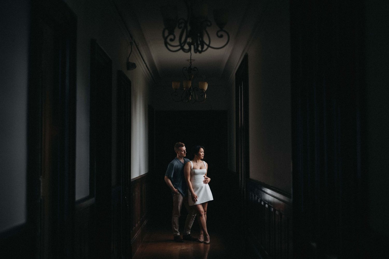 A couple holds hands walking through a dimly lit vintage hallway with chandelier overhead.