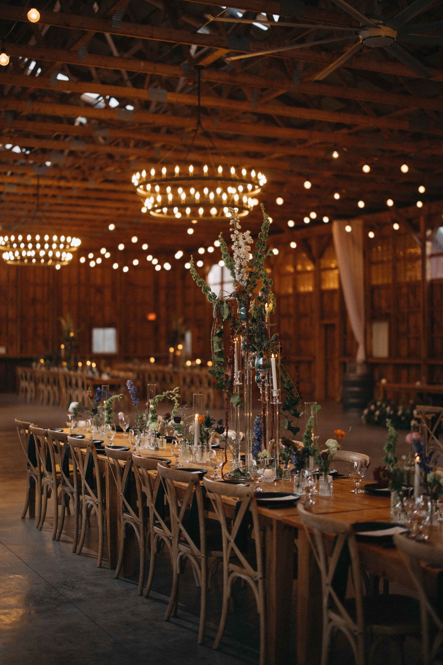 Rustic barn wedding reception with warm string lights, wooden tables, and tall floral centerpieces.