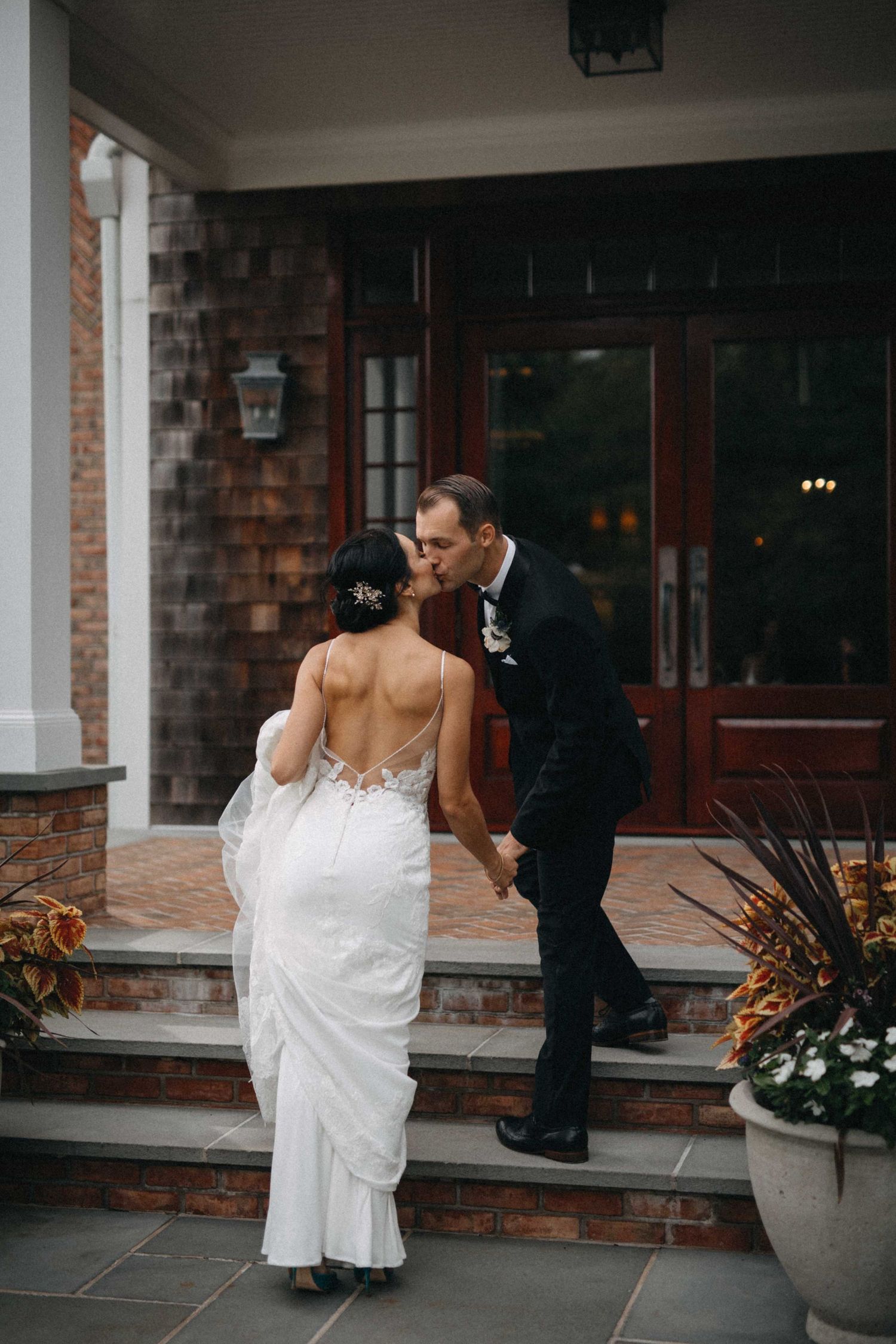 Bride and groom share a romantic kiss on the steps of a brick venue entrance while holding hands.