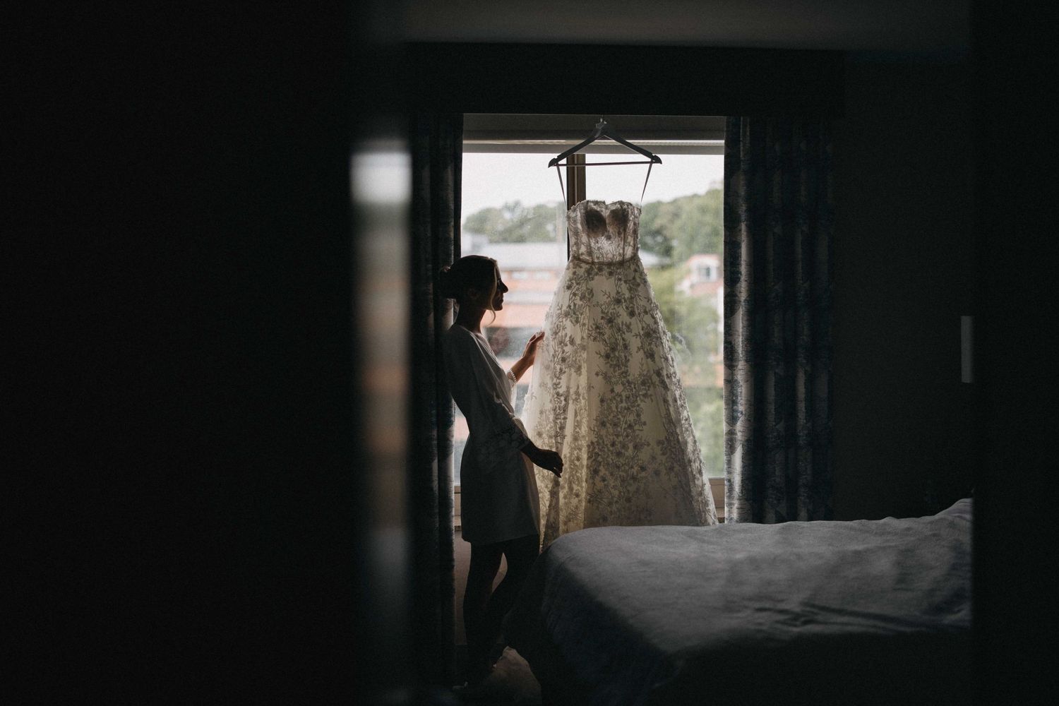 Silhouette of bride admiring lace wedding dress hanging in sunlit hotel room window.