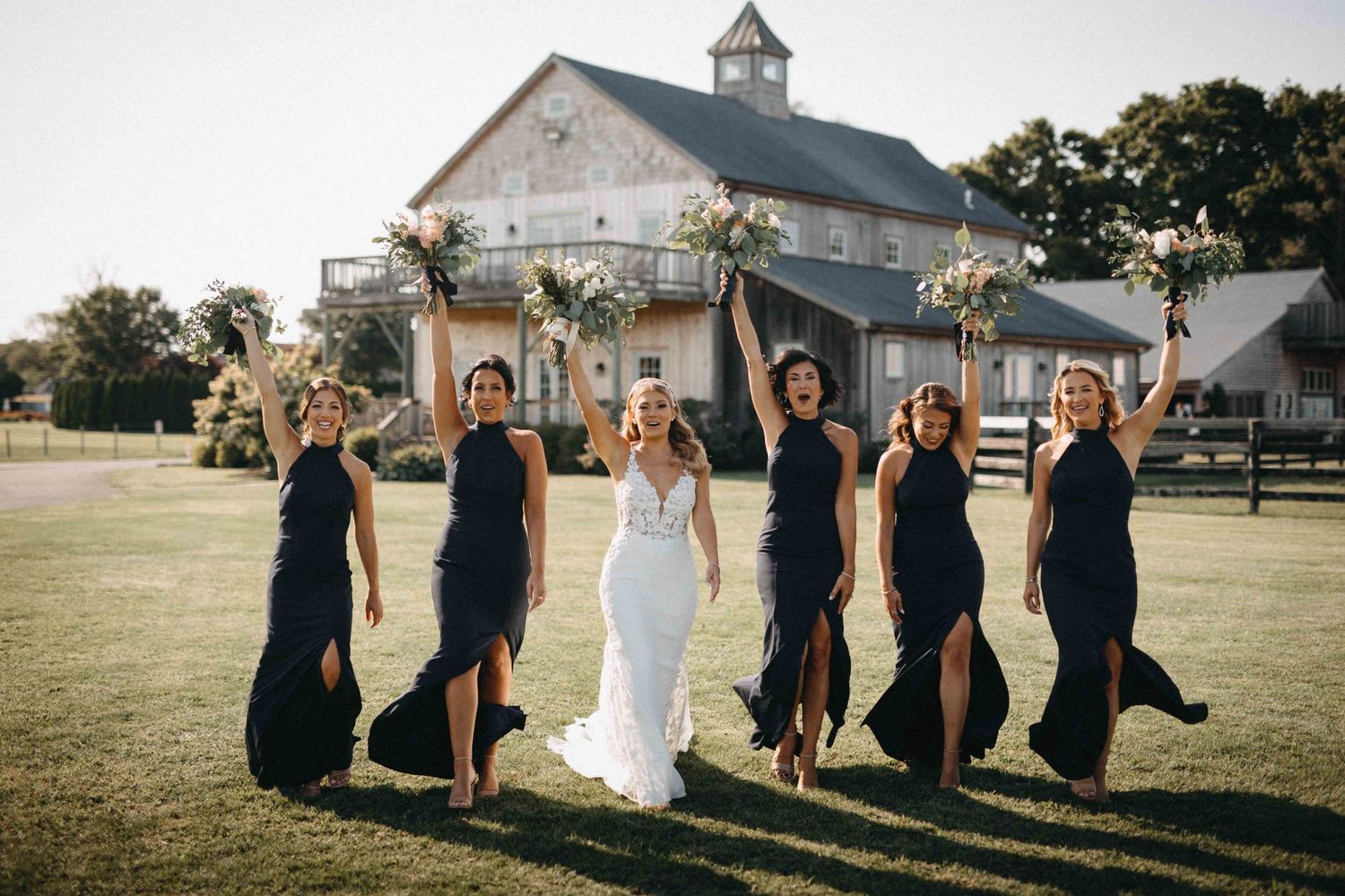 Bride in lace gown and bridesmaids in black dresses celebrating on green lawn with barn venue backdrop.