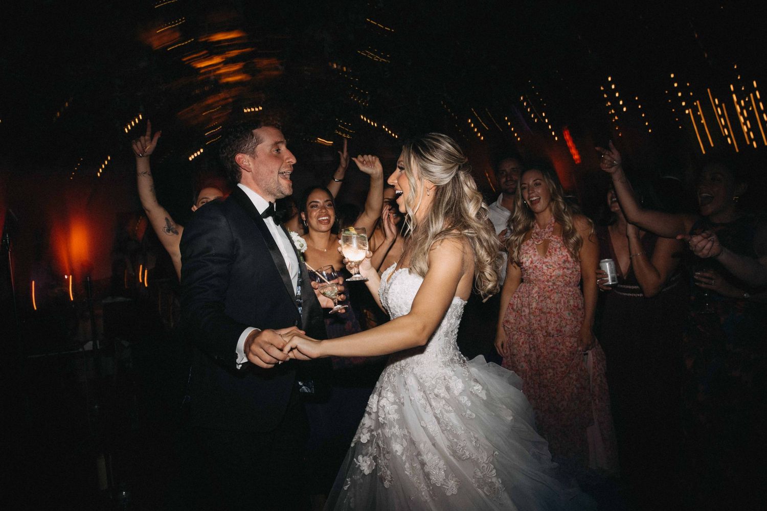 Bride in strapless white gown and groom in tuxedo dancing joyfully with guests at lively wedding reception.