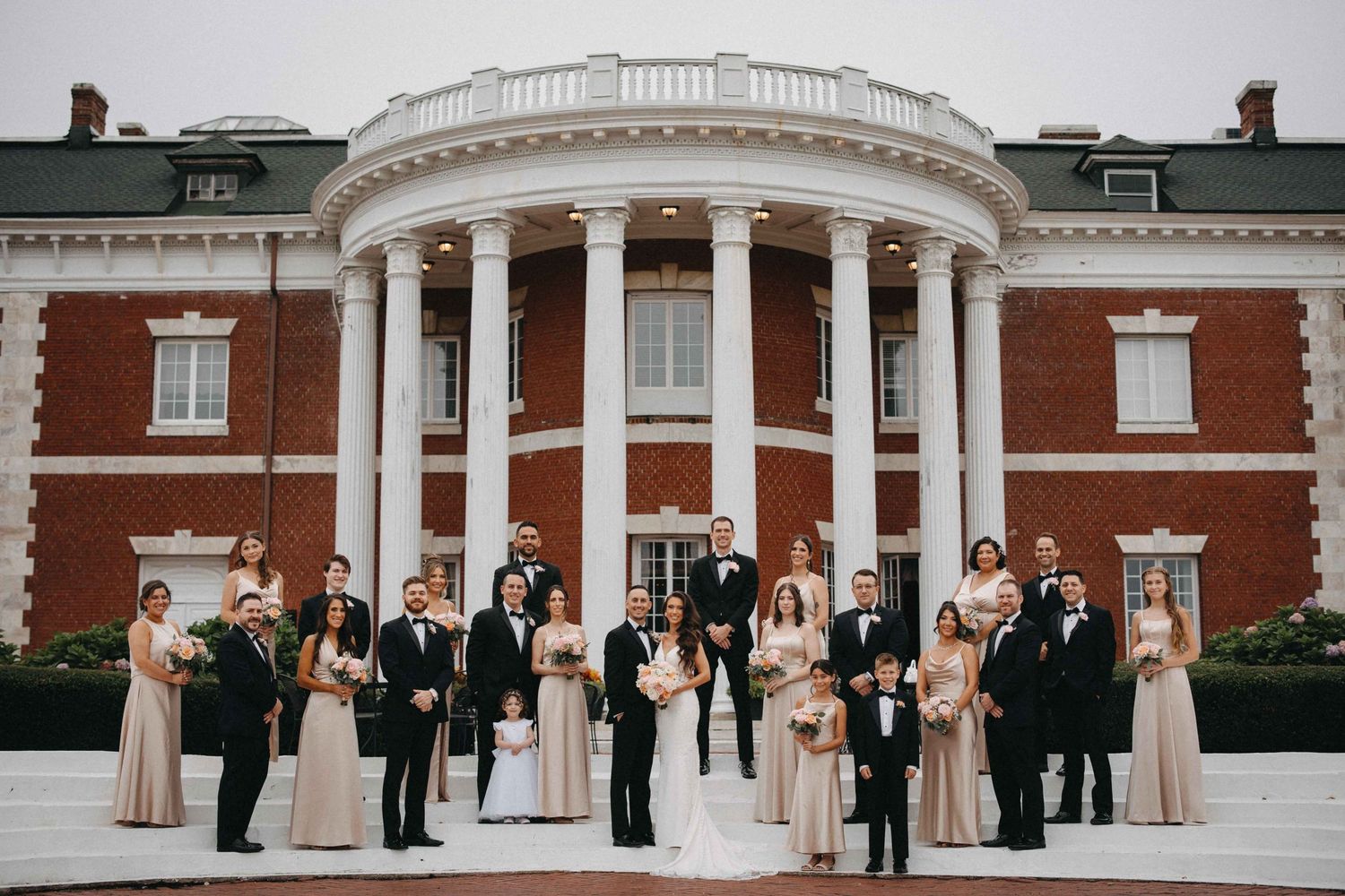 Large wedding party poses before grand red brick colonial mansion with white rotunda entrance.