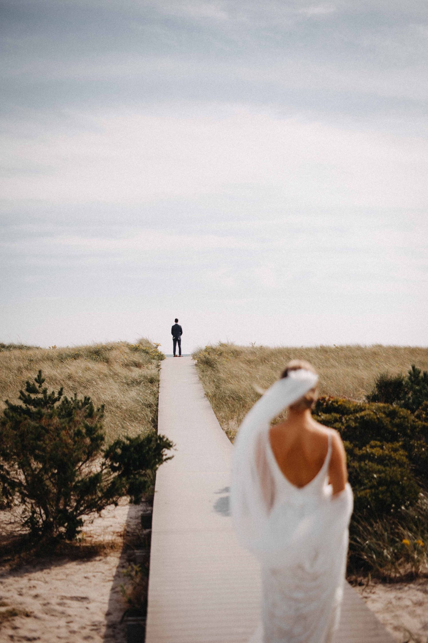 Bride in backless wedding gown walks boardwalk toward groom standing at sandy dune horizon.