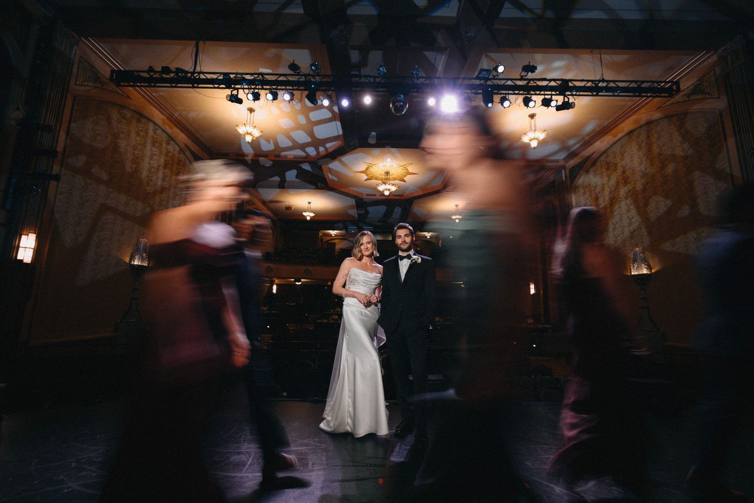 Bride and groom standing together surrounded by blurred dancing guests in elegant reception hall.