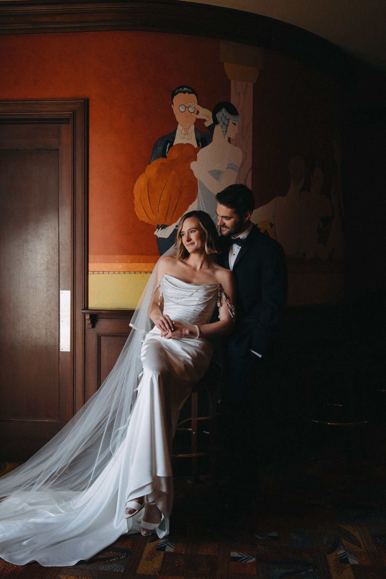 Bride and groom pose elegantly beneath a vintage mural in a warmly lit wedding venue.