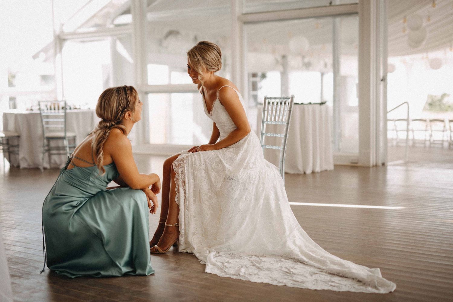 Bridesmaid in sage green dress helping bride with her lace gown in a bright venue.