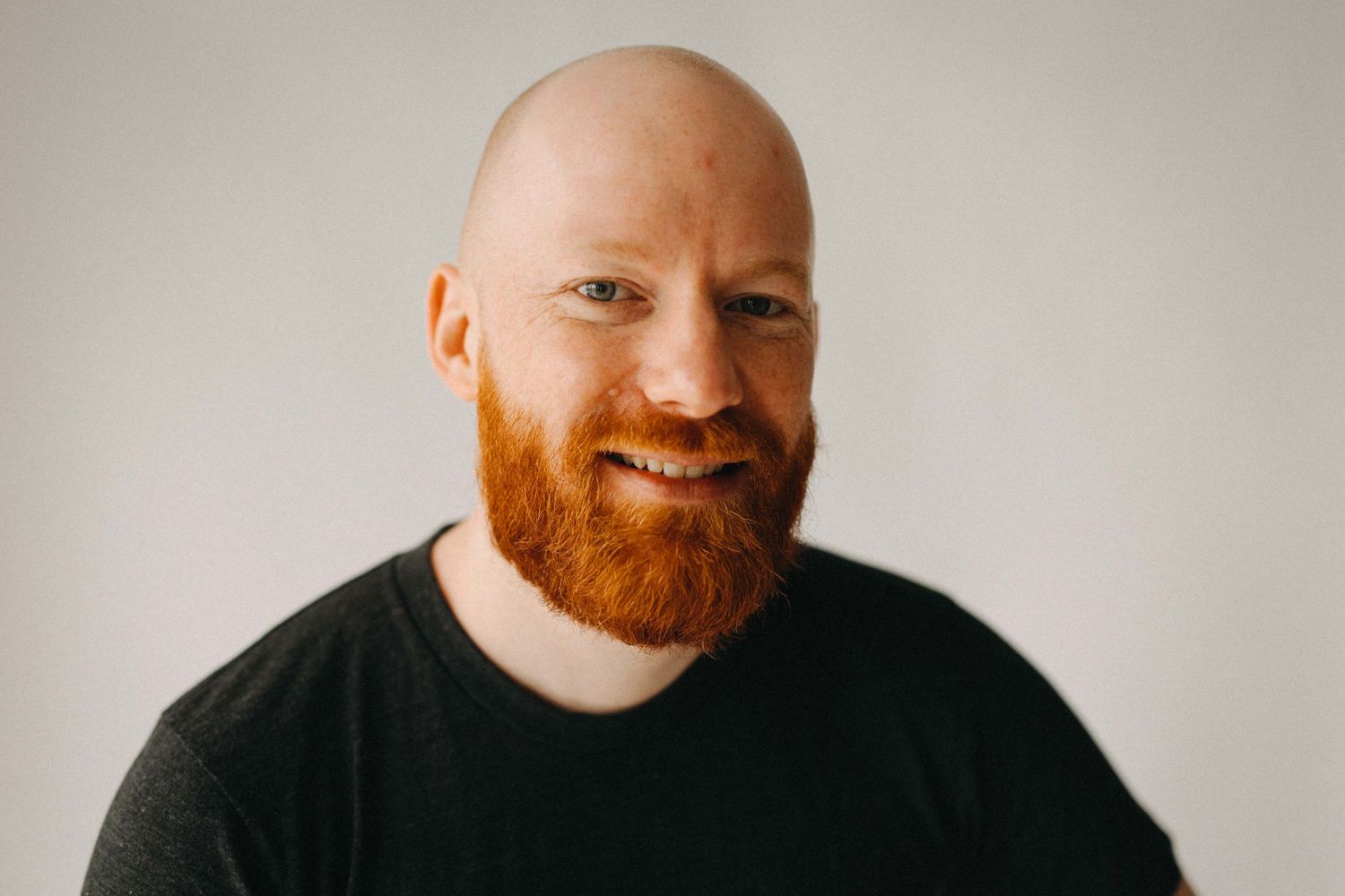 Bald man with red beard smiling warmly wearing black t-shirt against light gray background.