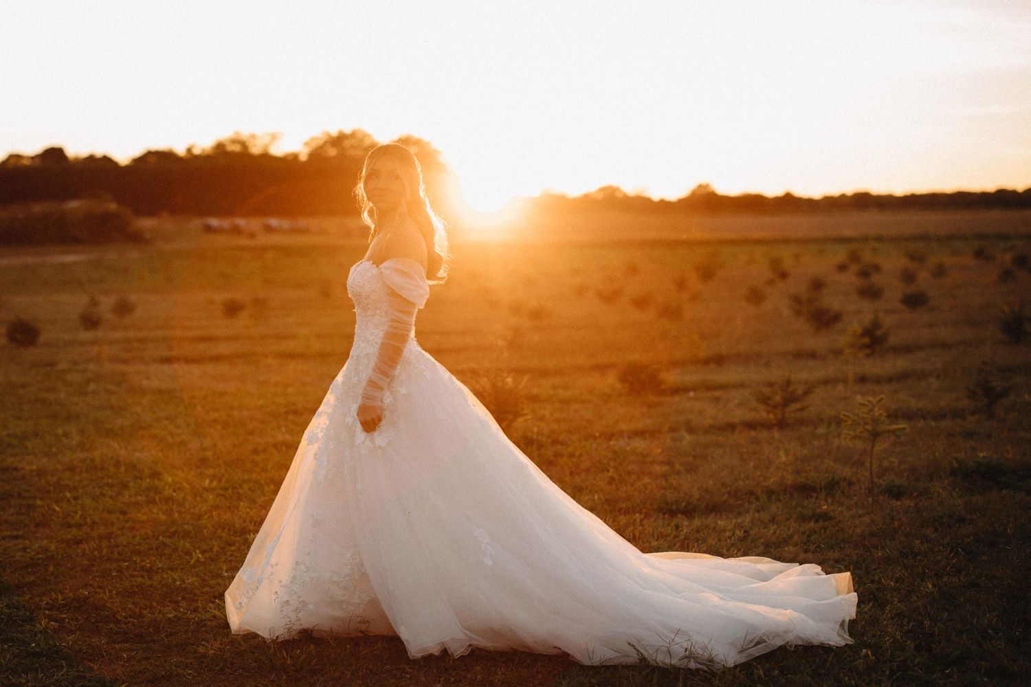 Bride in an elegant off-shoulder white ball gown with cathedral train posing in a sunlit field at golden hour.