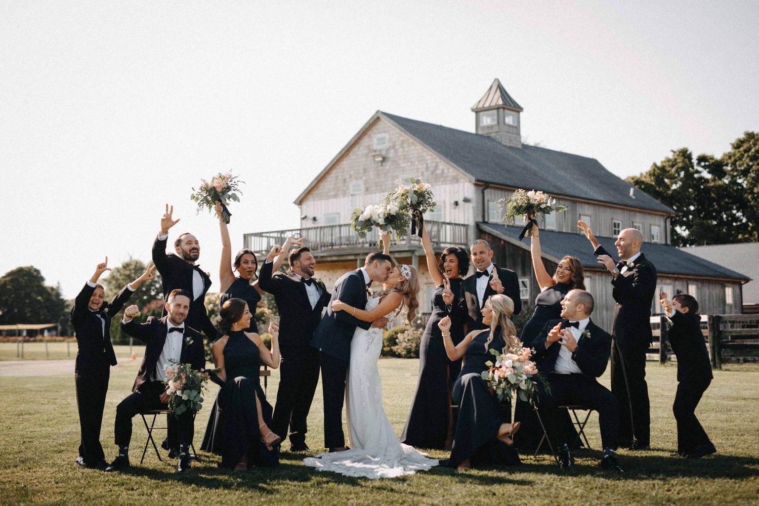 Wedding party celebrating with raised bouquets in front of rustic barn venue.