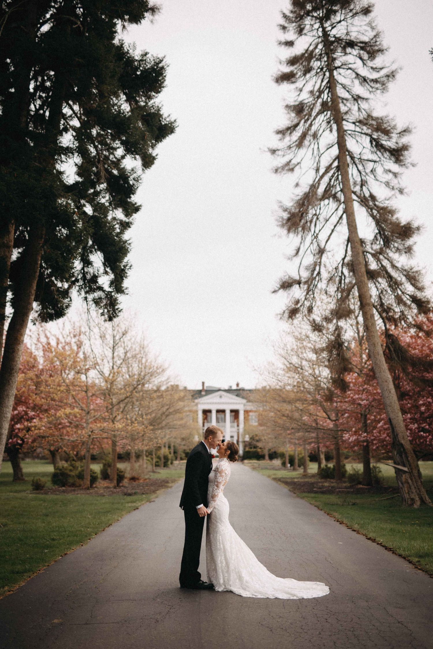 Bride and groom share romantic kiss on tree-lined path before grand white colonial mansion.