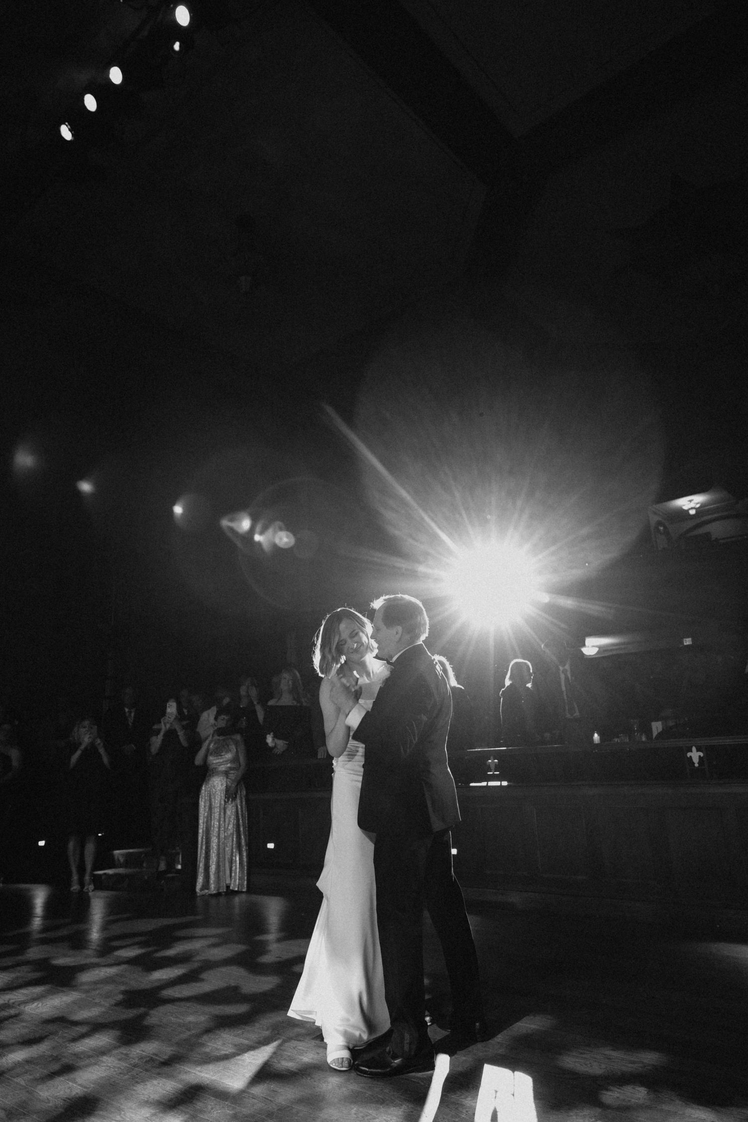 Black and white photo of a couple sharing their first dance at a wedding reception under a bright spotlight.