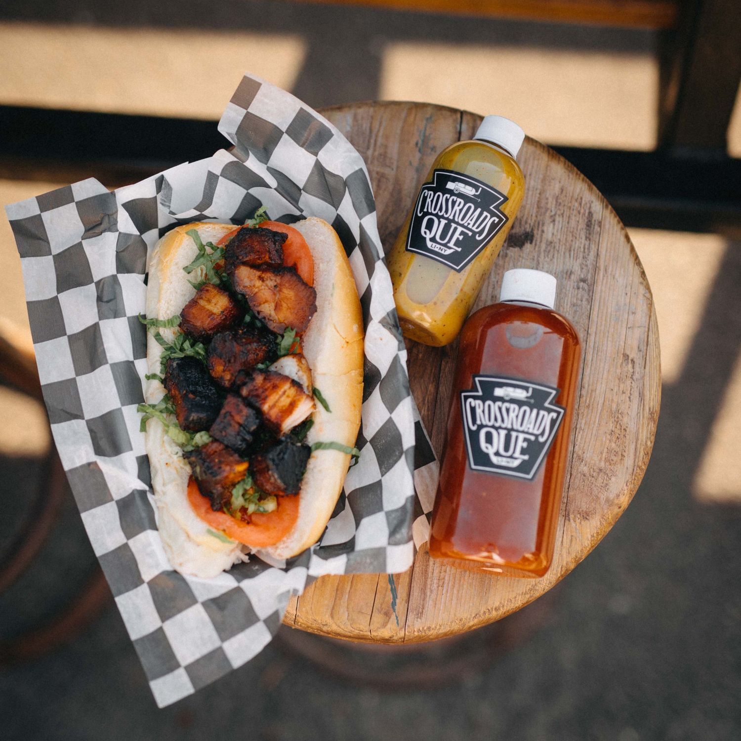 BBQ sandwich with charred meat on checkered paper beside two Crossroads Que sauce bottles on a wooden table.