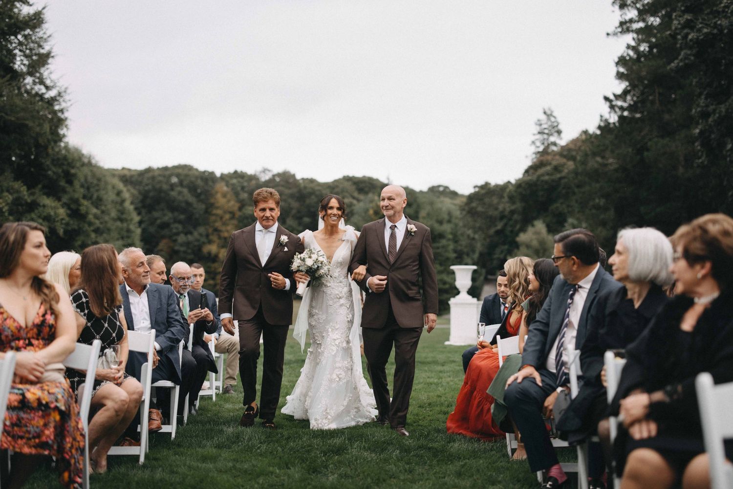 Bride walks down outdoor aisle escorted by two men at a lush garden wedding ceremony with white chairs and guests.